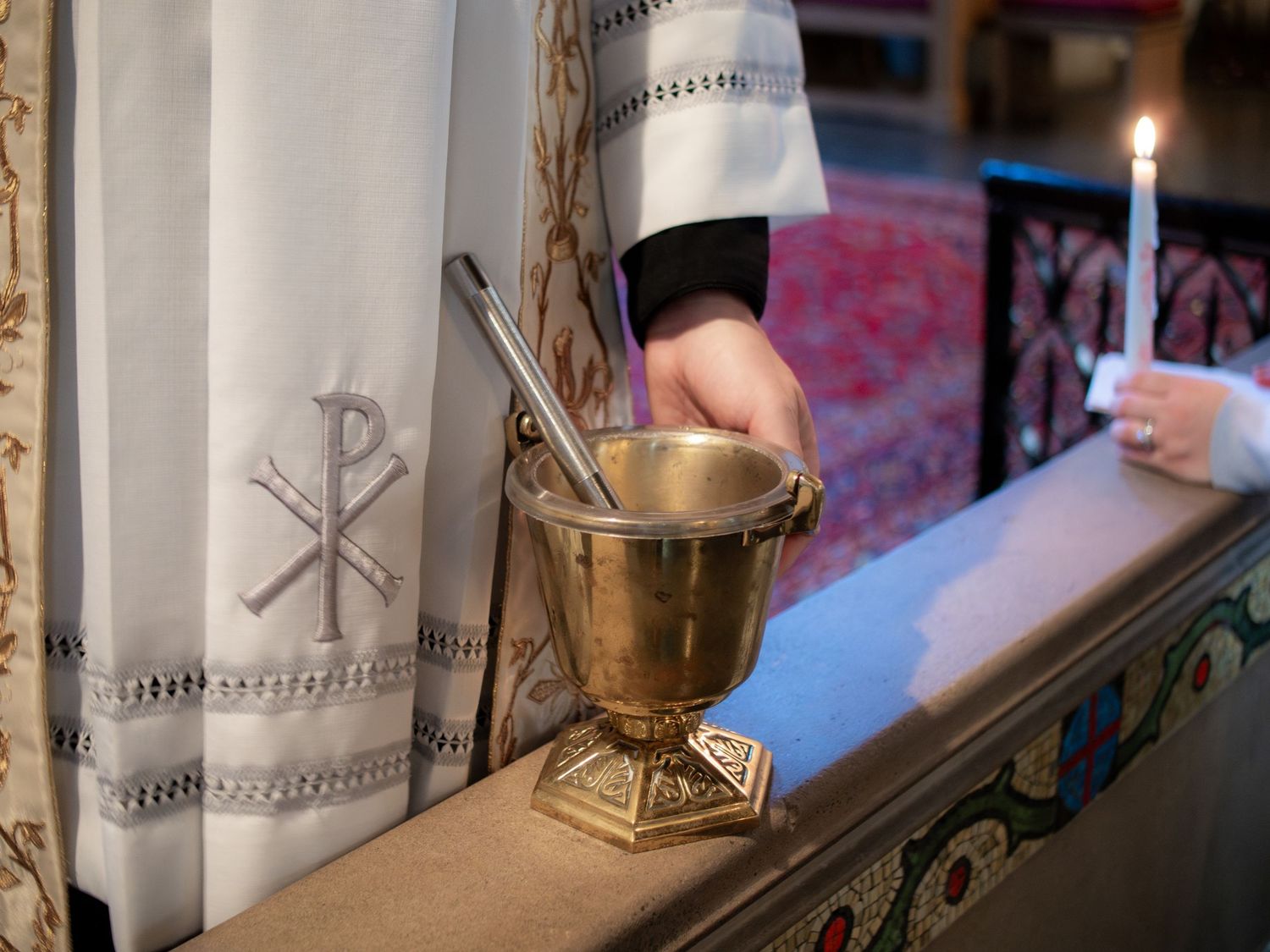 Brass holy water bucket with aspergillum beside Chi Rho embroidered white vestment in church.