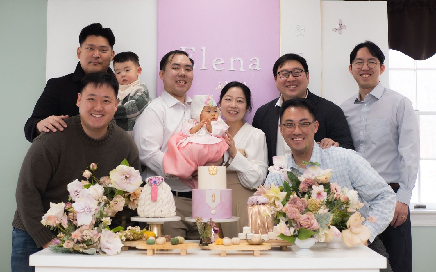Group of people celebrating a baby shower with floral arrangements, cake, and a baby dressed in pink.