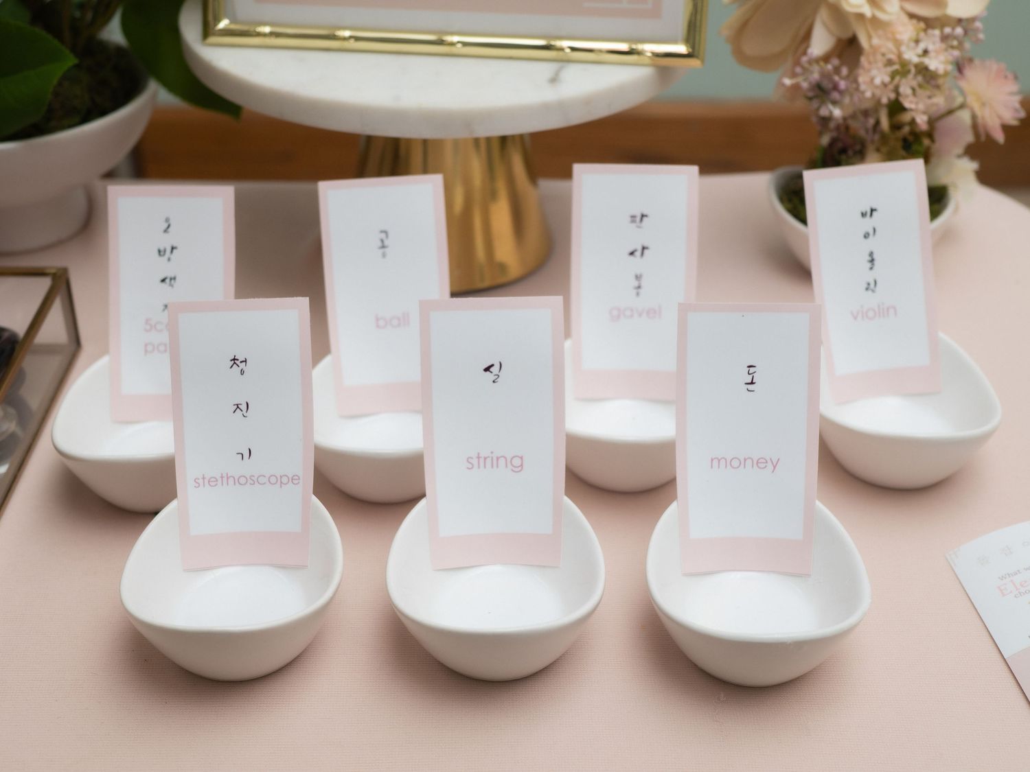White ceramic bowls with pink cards displaying Korean characters for a traditional doljabi fortune-telling ceremony.