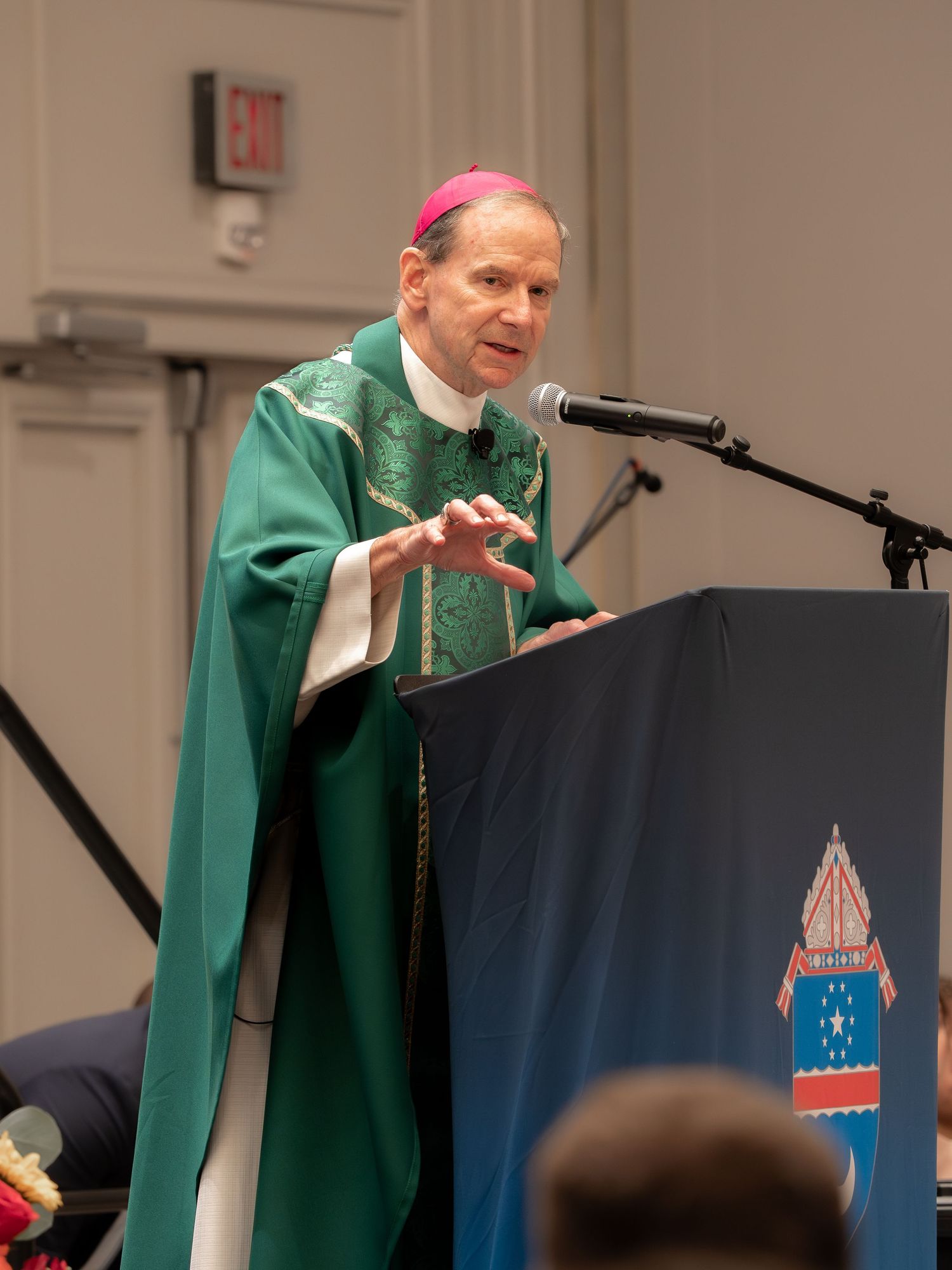 Michael F. Burbidge, Bishop of the Diocese of Arlington, wearing green vestments and mitre speaking passionately at a blue decorated podium.