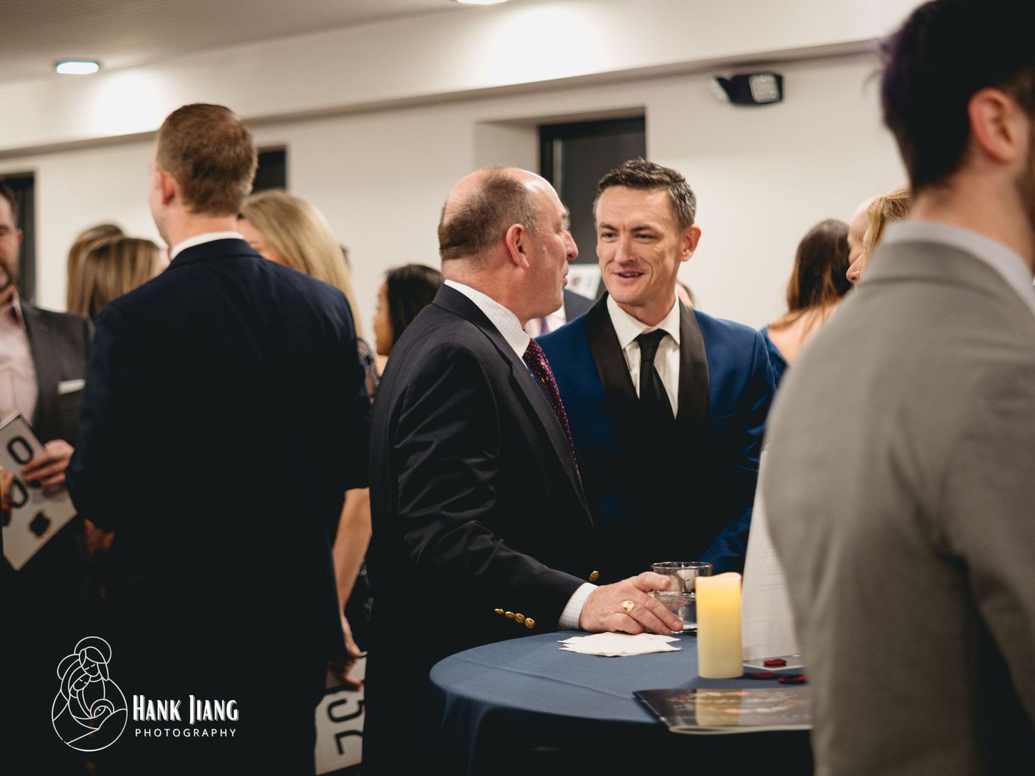 Two men in suits shaking hands and smiling at a formal networking event or cocktail reception.