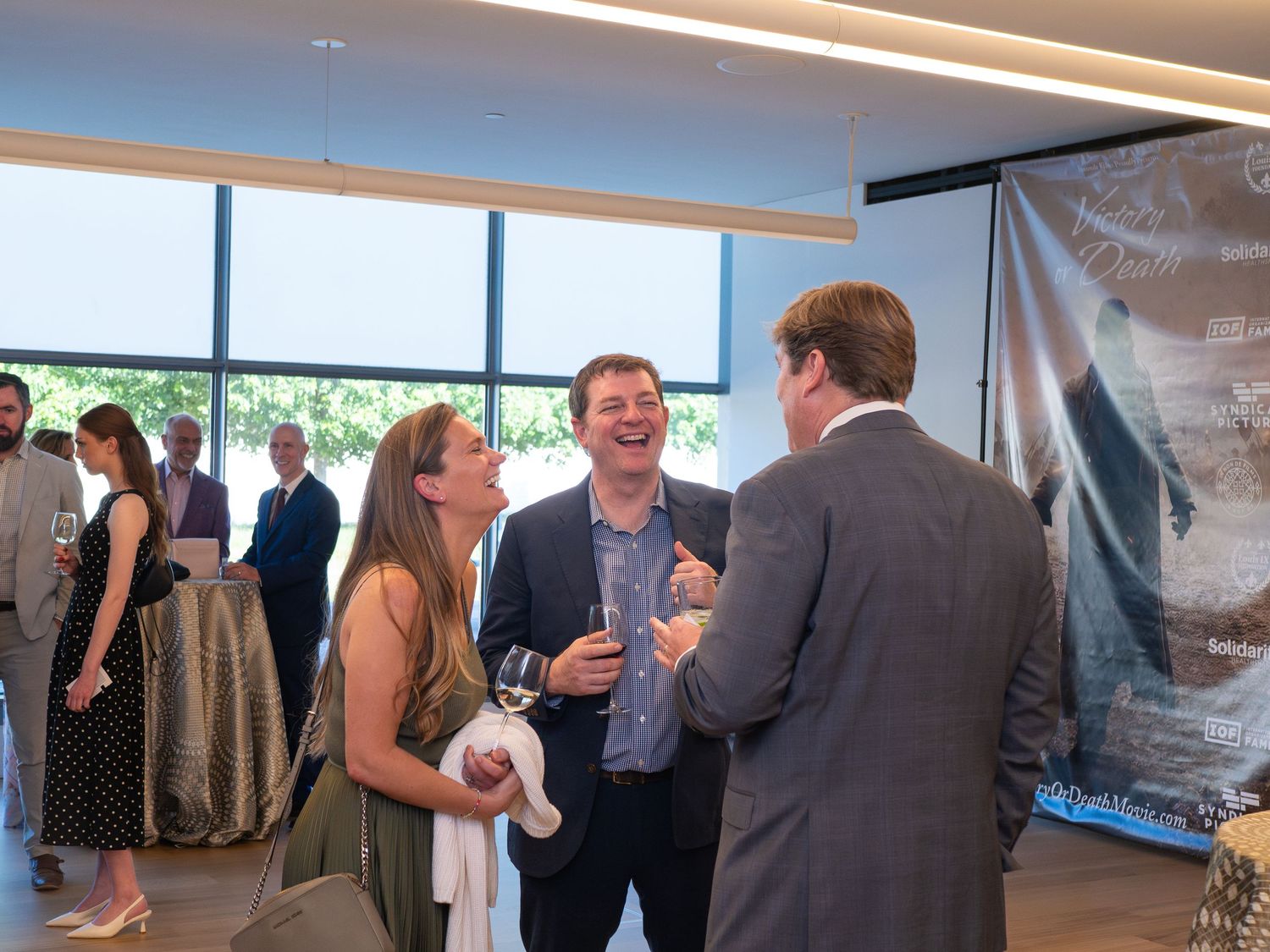 Three professionals networking and laughing at an indoor corporate event with large windows and a banner display.