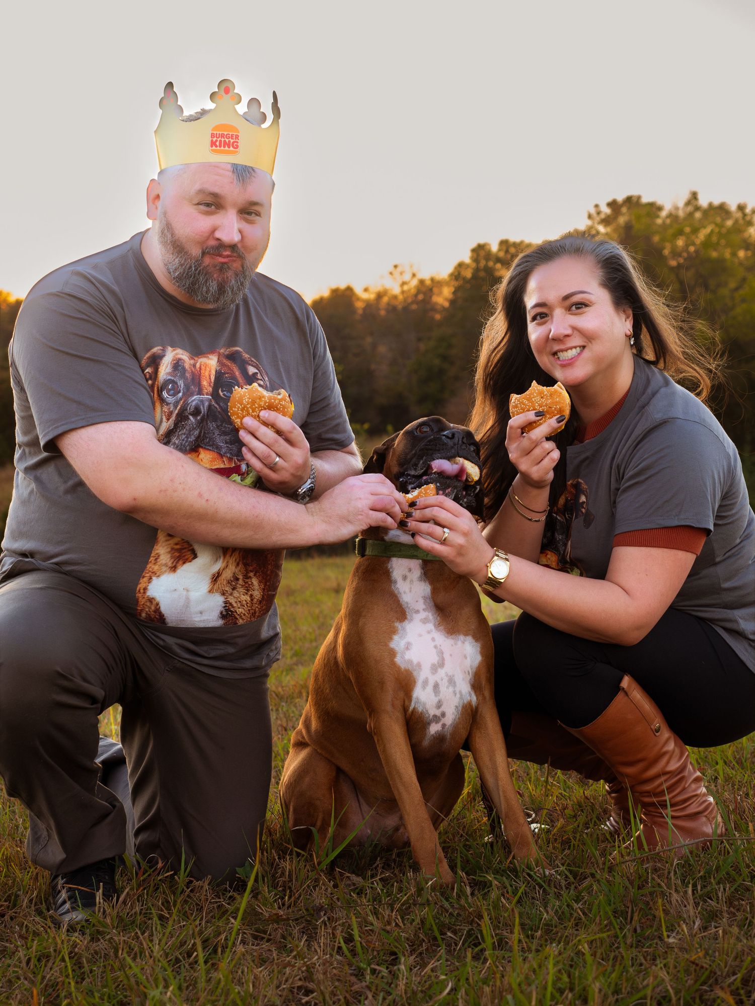 Couple wearing Burger King crowns feeding burgers to their boxer dog outdoors at golden hour.