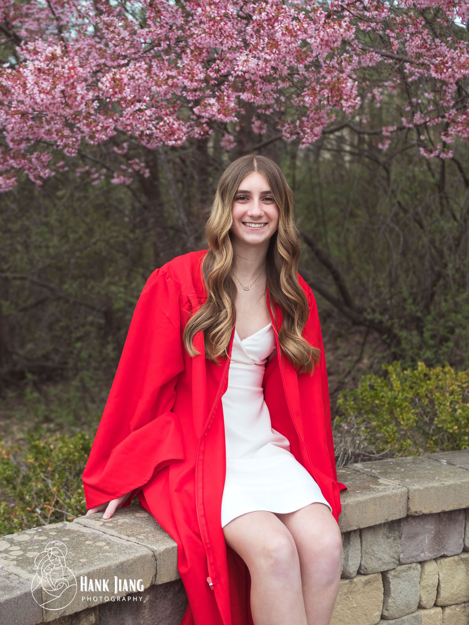 Smiling graduate in red cap and gown sits on stone wall with pink cherry blossoms in spring outdoor setting.