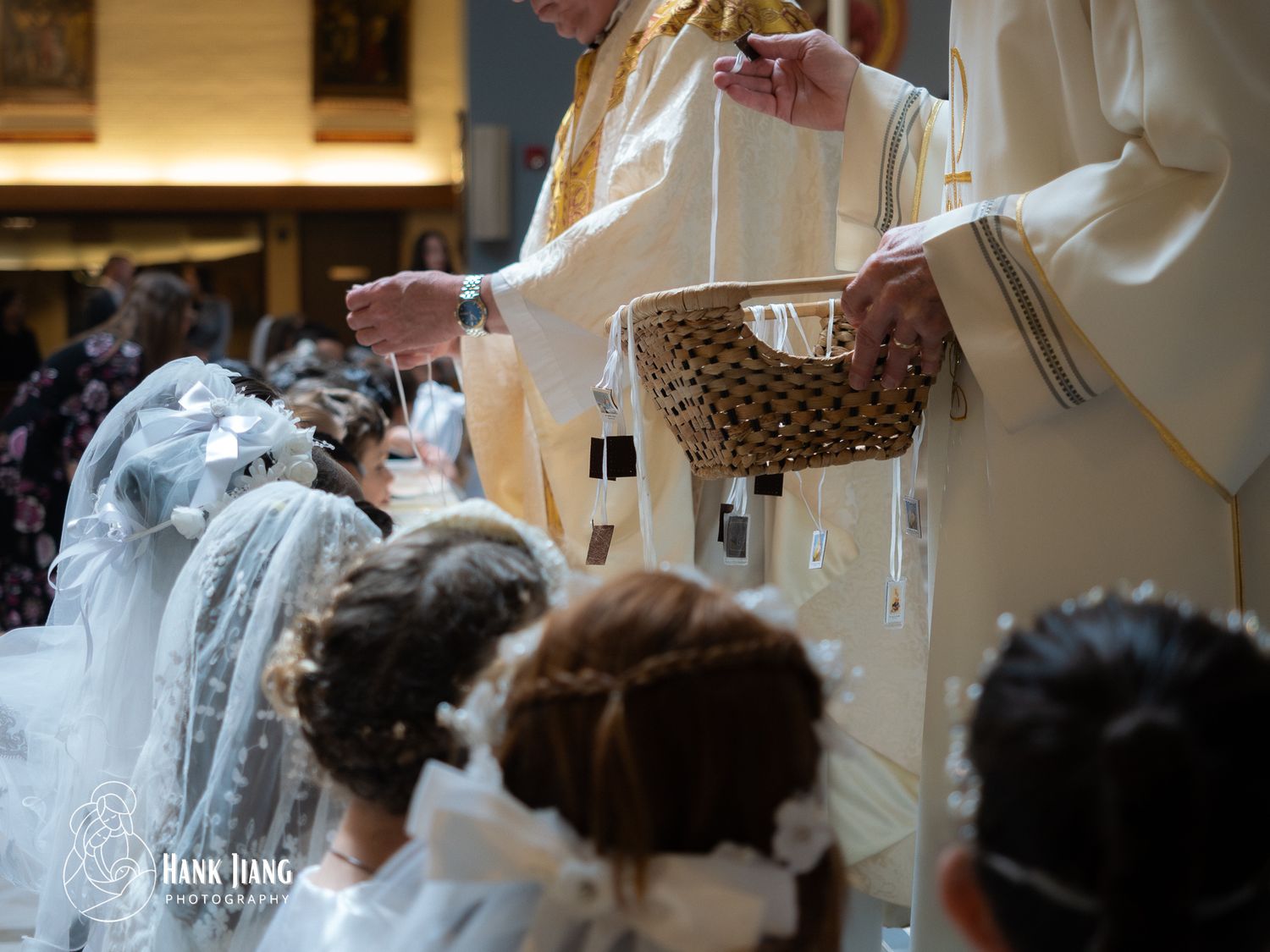 Priest performing First Communion ceremony with young girls in white veils kneeling in a church.