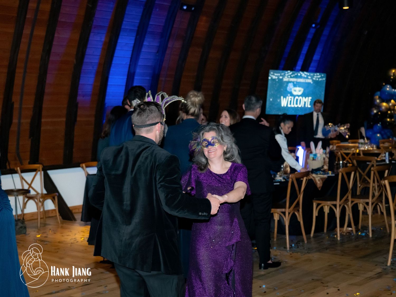 Guests in formal attire dance and mingle at an elegant masquerade ball with welcome signage.