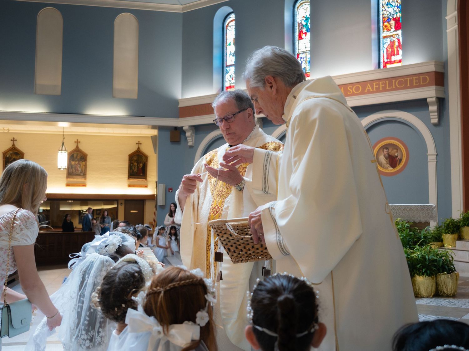 Two priests administer First Communion to young girls in white veils inside a decorated Catholic church.