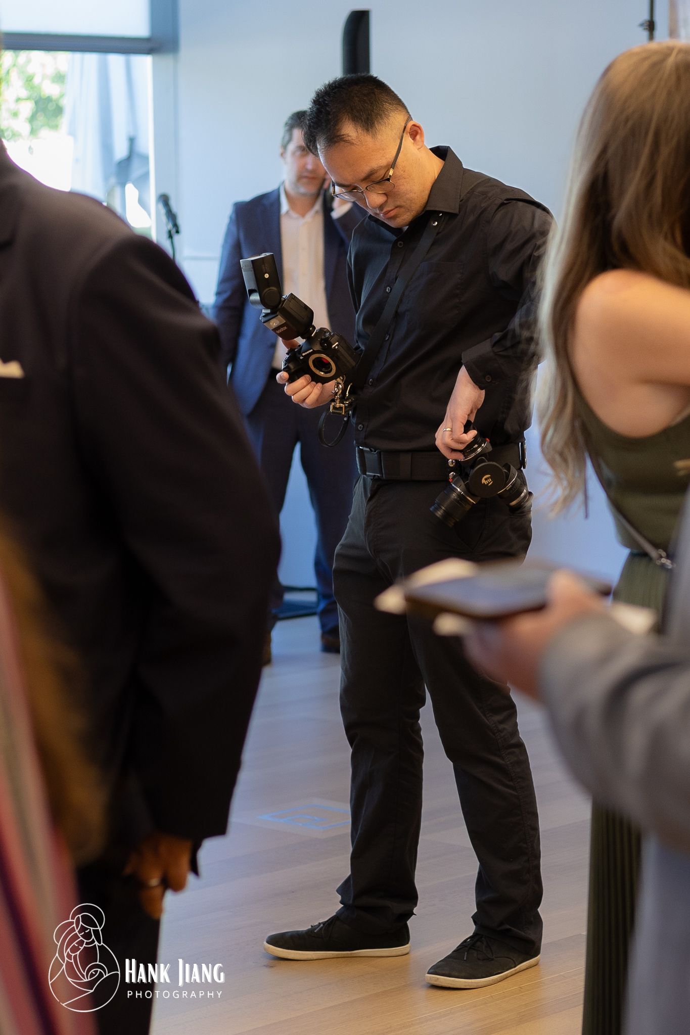 Photographer in black outfit holding two cameras at an indoor event, with guests visible in the background.