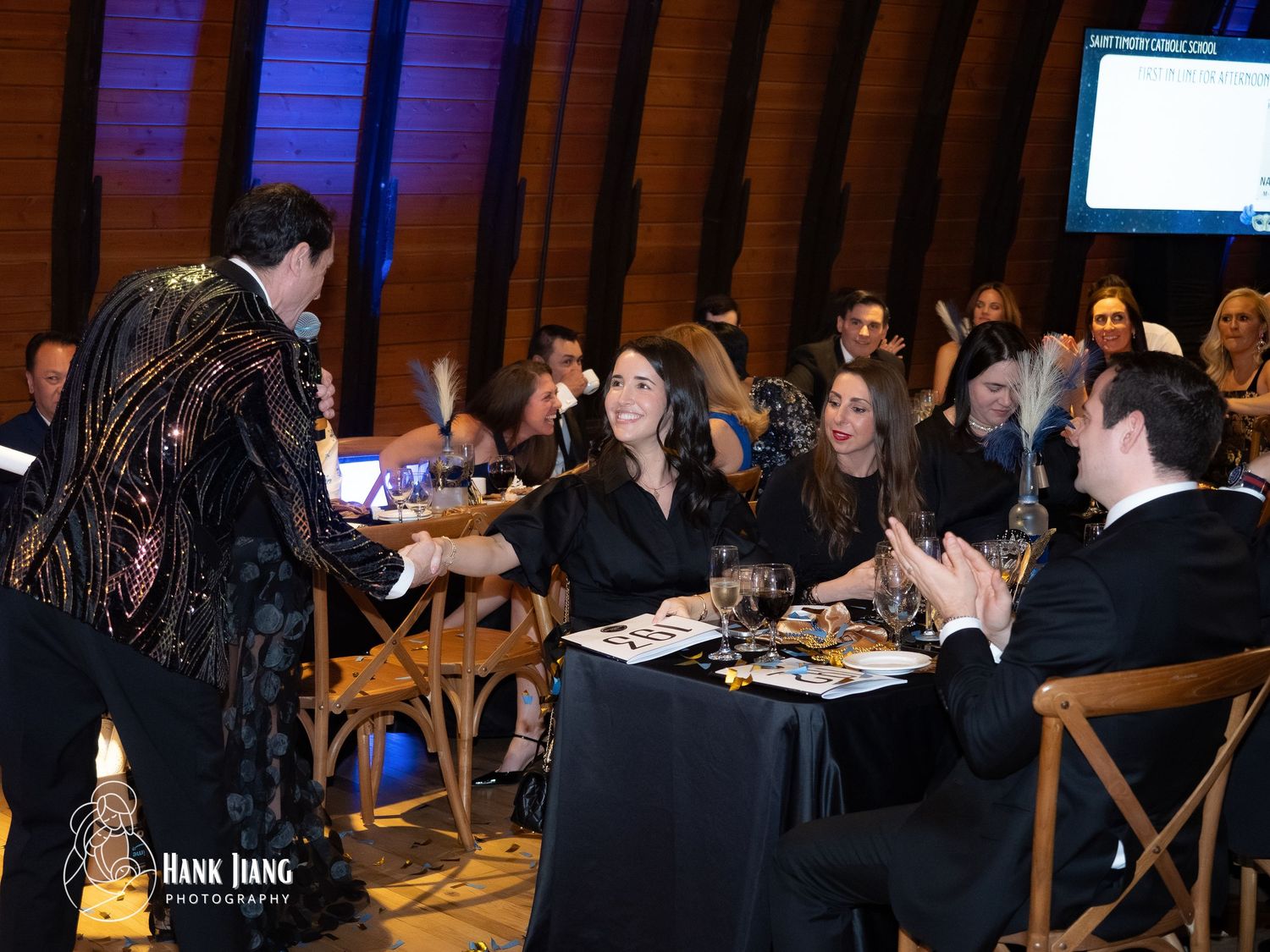 Elegant gala event with guests in formal attire seated at tables, as a performer in a sequined jacket greets attendees.
