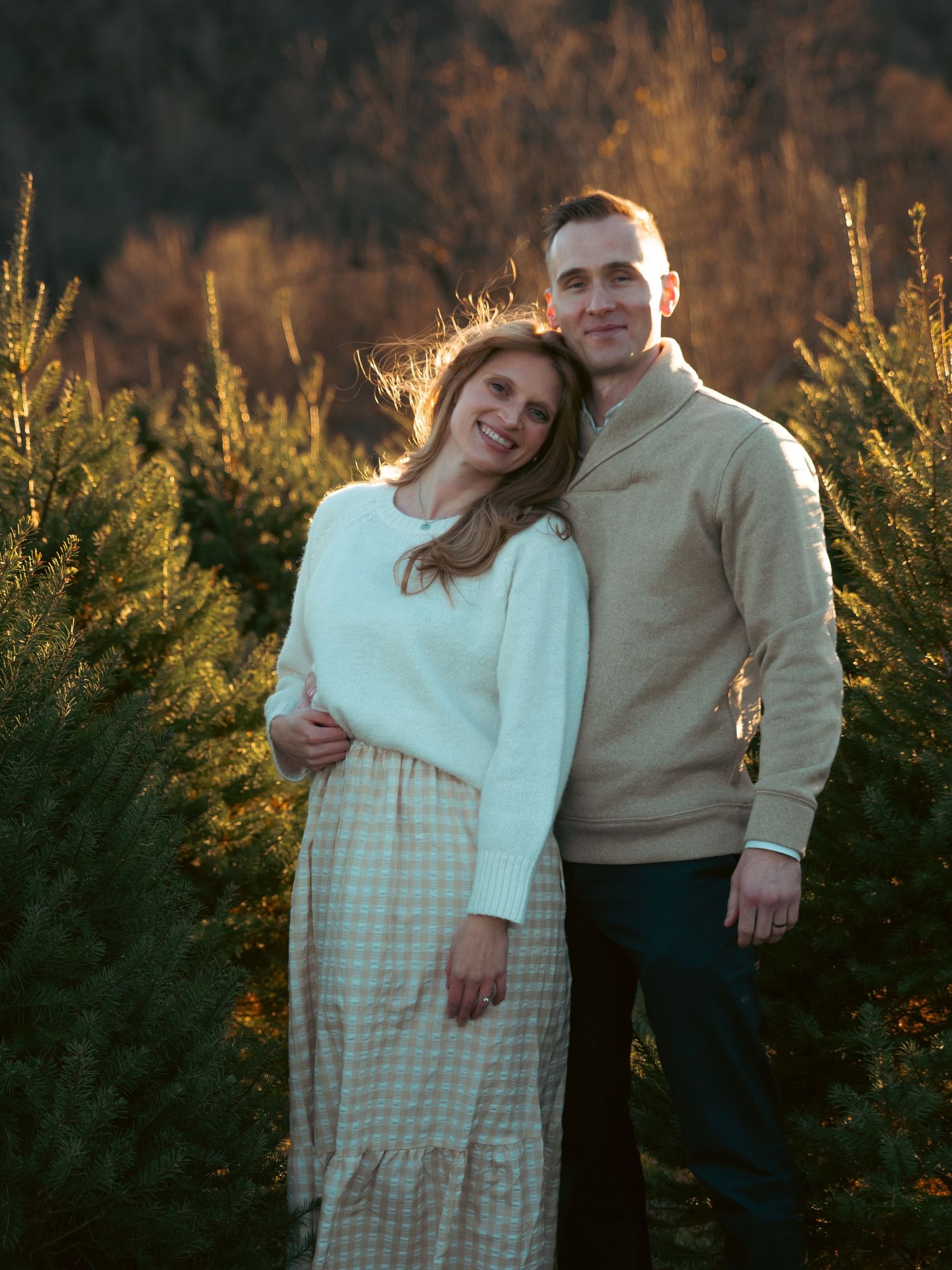 Smiling couple poses together at golden hour among Christmas tree farm rows.