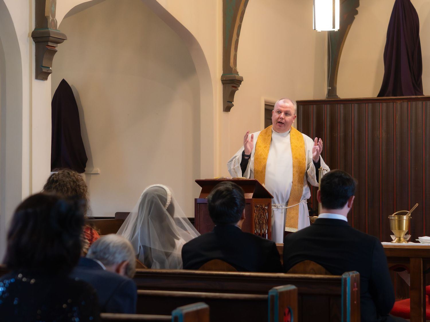 A priest in white and gold vestments addresses wedding guests seated in wooden church pews during a ceremony.