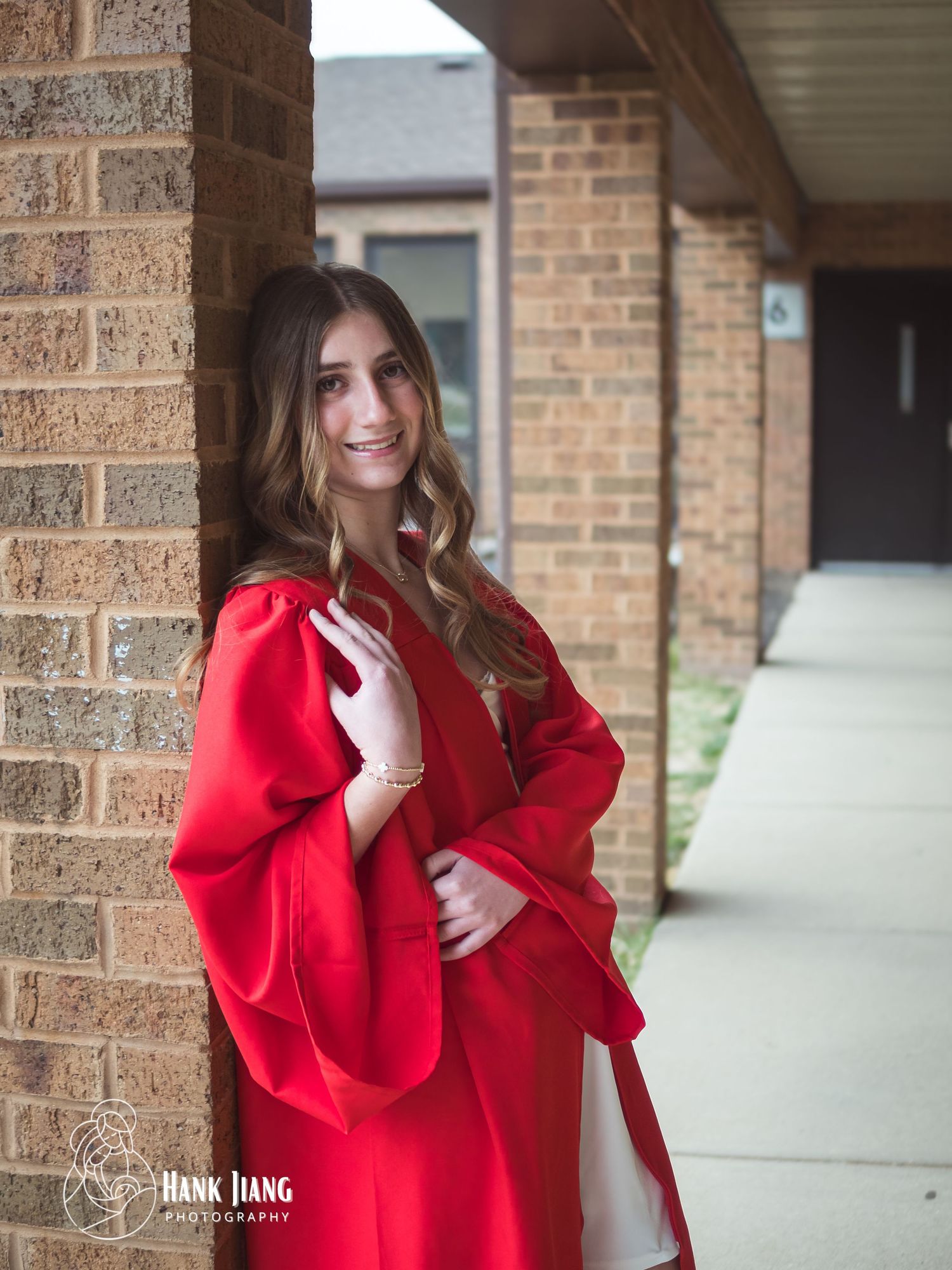 Young woman in red graduation gown leaning against brick wall, smiling for senior portrait photo.