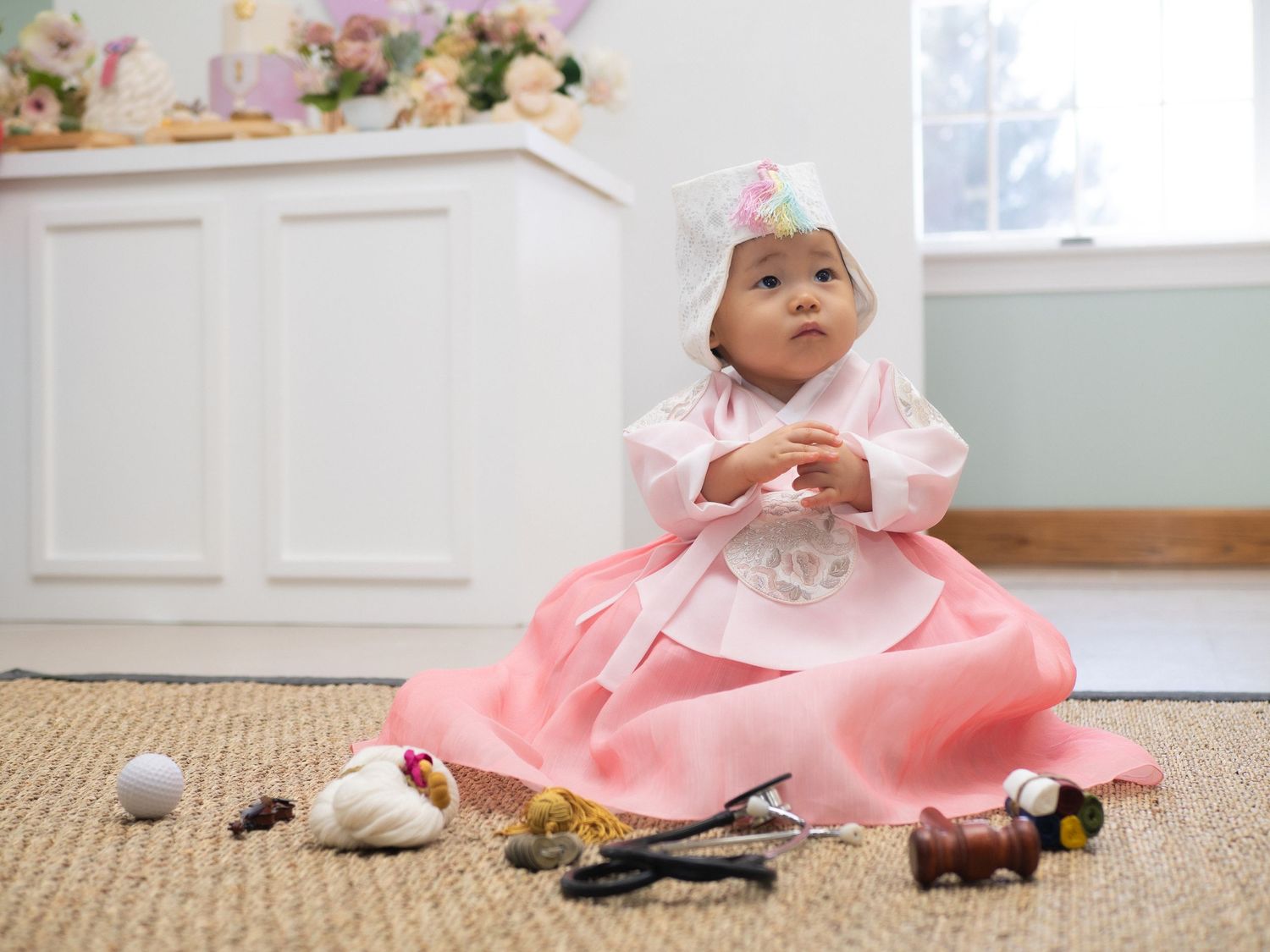 Baby in pink Korean hanbok dress sitting on floor surrounded by traditional doljabi objects during first birthday celebration.