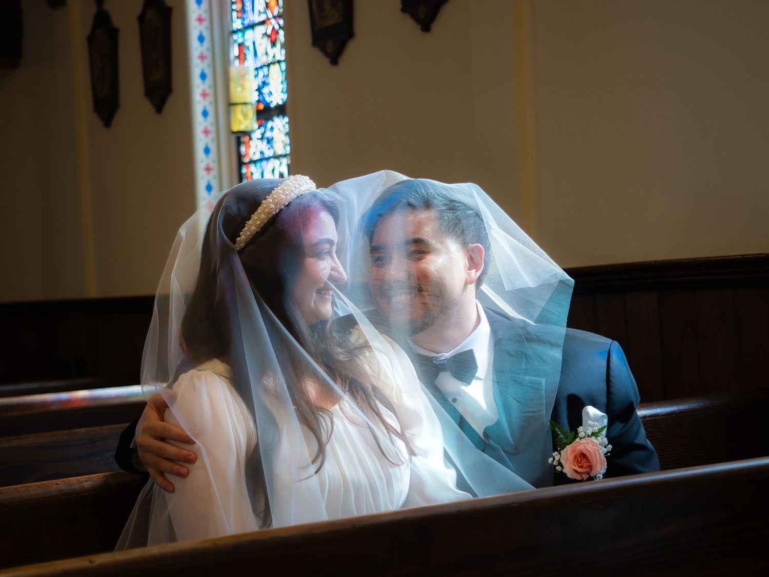 Newlywed couple sharing an intimate moment under a wedding veil in a church pew with stained glass windows.