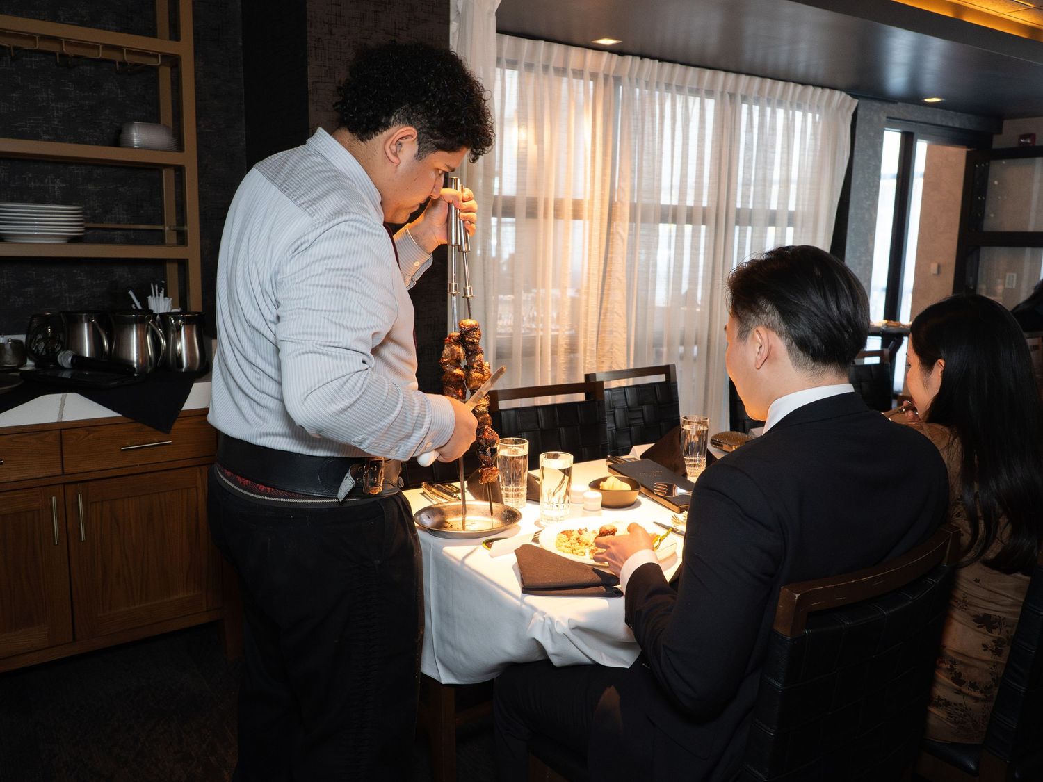 Waiter serving food to couple dining at elegant restaurant table with white tablecloth and warm lighting.
