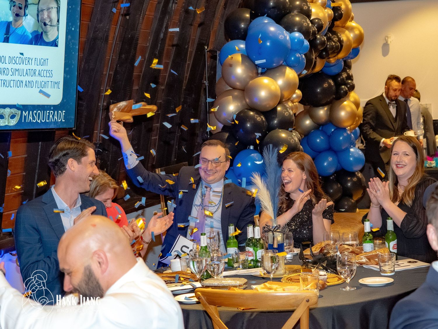 Guests celebrate at a masquerade gala with blue and gold balloons, confetti, and festive table settings.