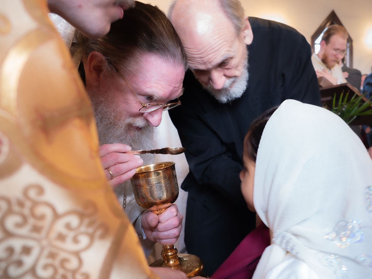 Orthodox priest administering communion chalice to young girl during church service.