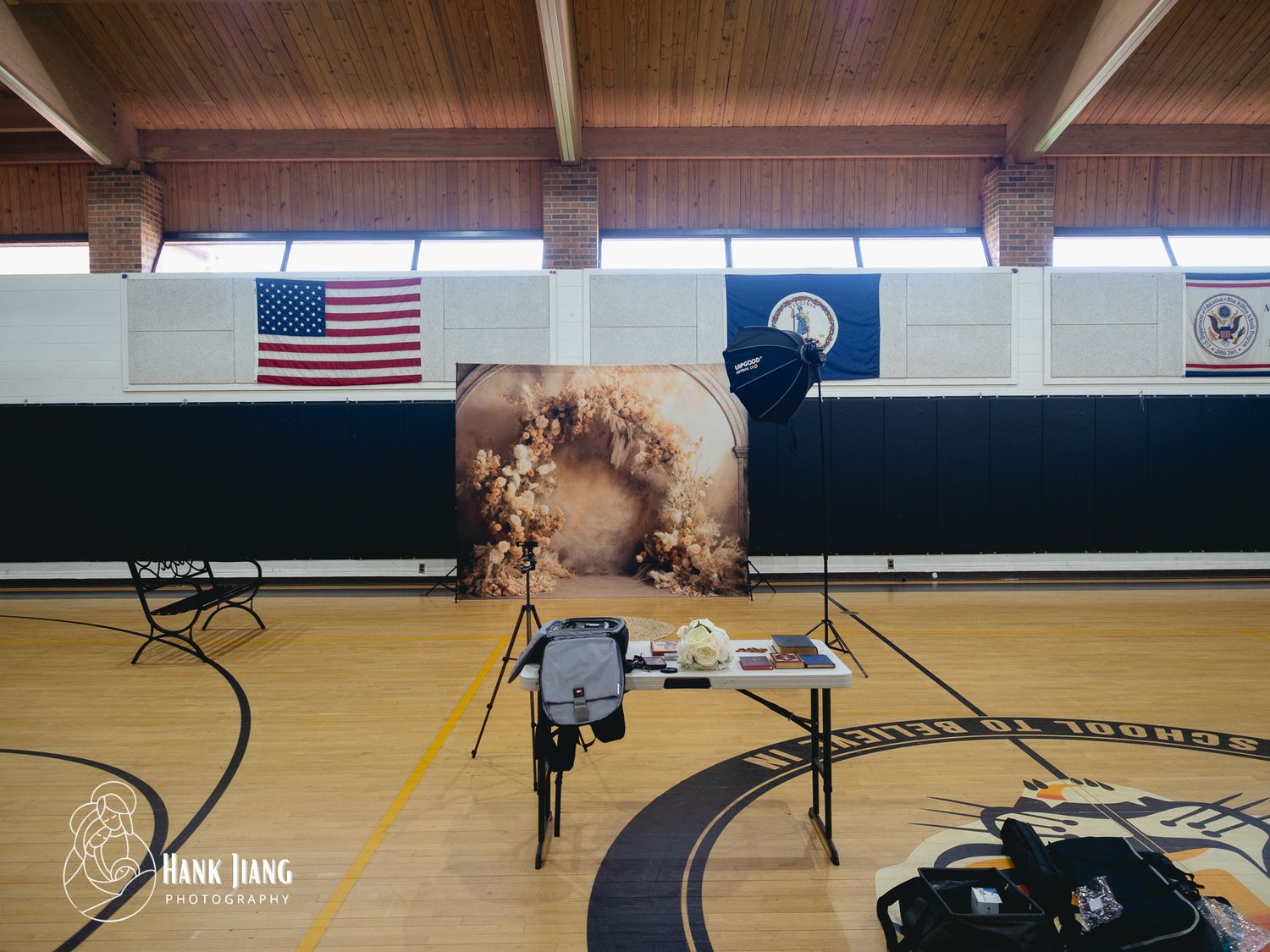 Photography studio setup in a school gymnasium with backdrop, lighting equipment, and American flag on the wall.
