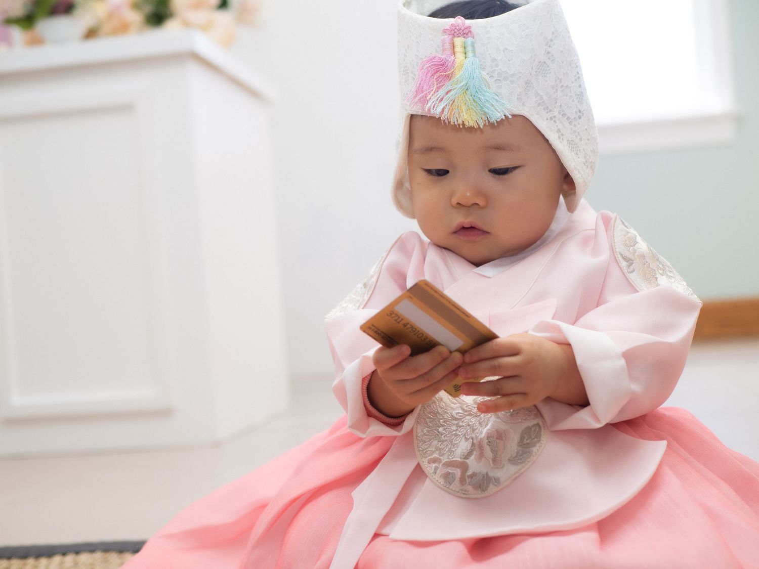 Baby in pink Korean hanbok and white gat hat holds a wooden book during a traditional dol first birthday celebration.