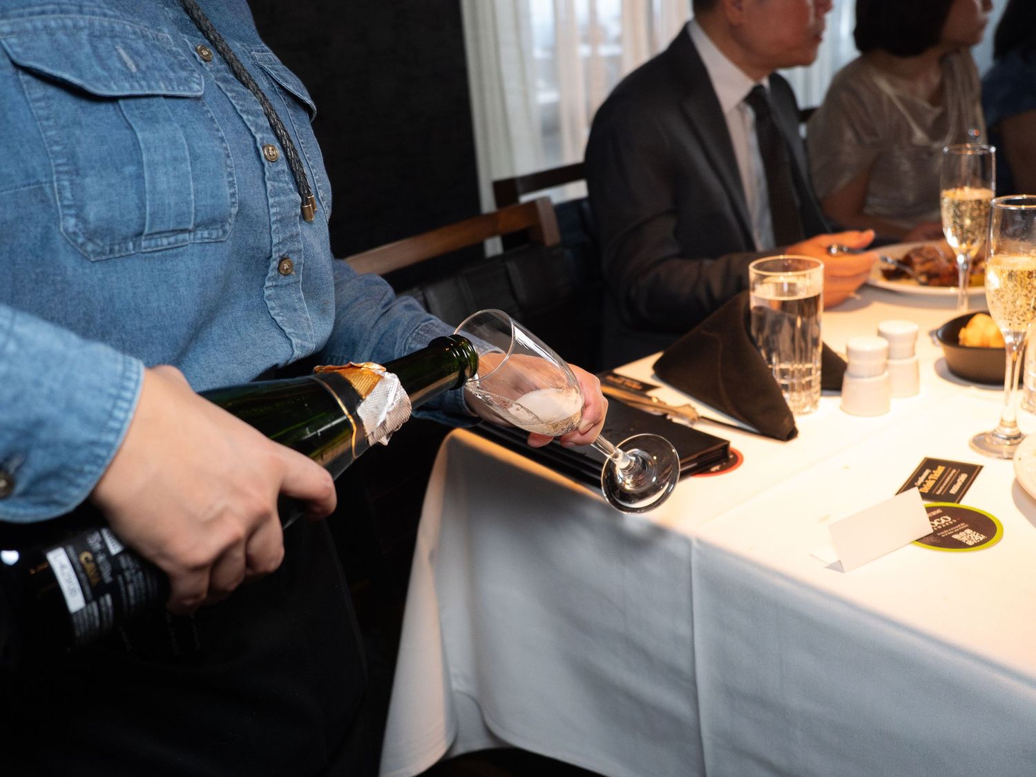 Server pouring champagne into a glass at an elegant restaurant dining table with guests in the background.