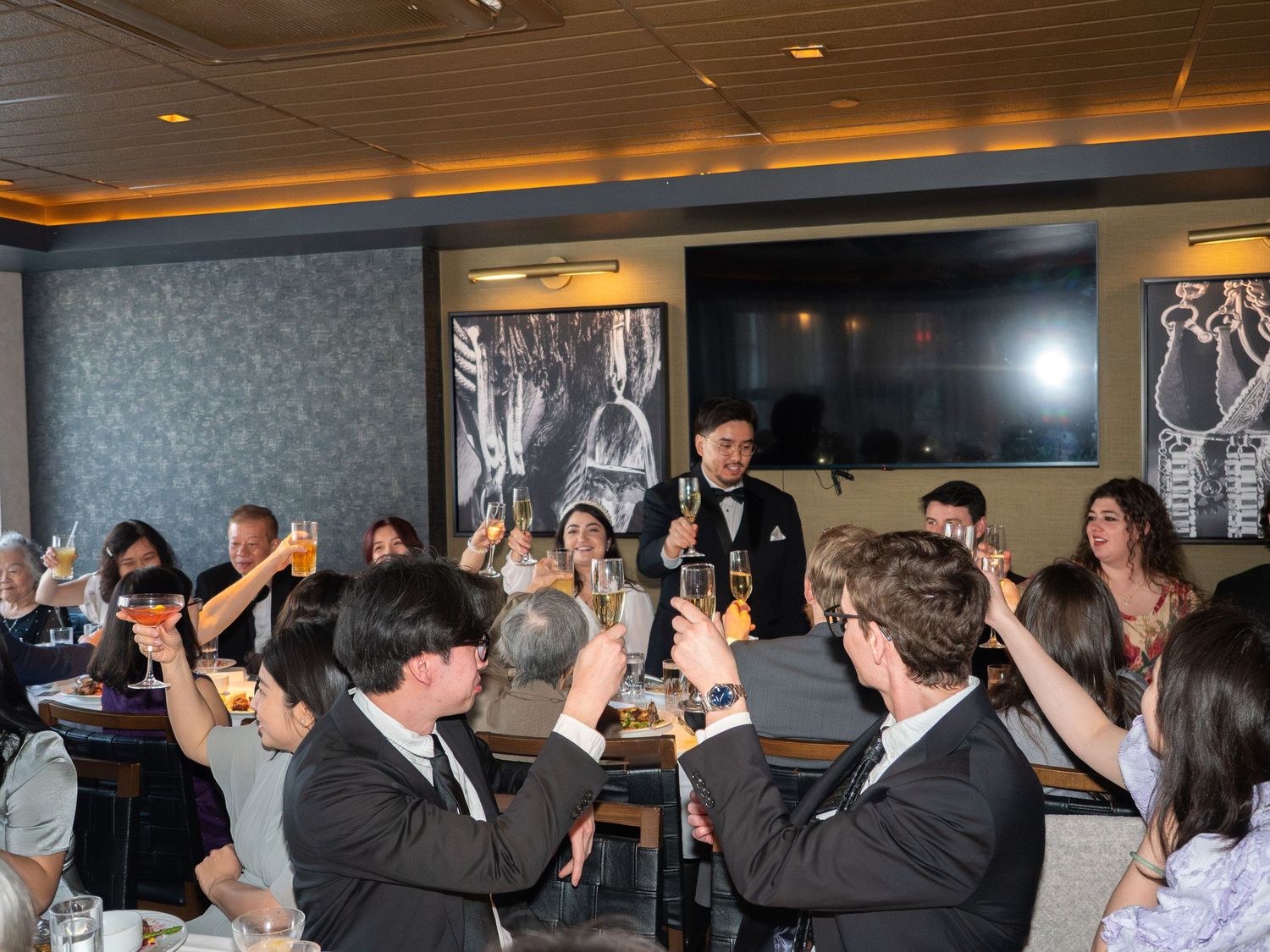 Guests raising glasses in a toast at an elegant restaurant celebration with formal attire and warm lighting.