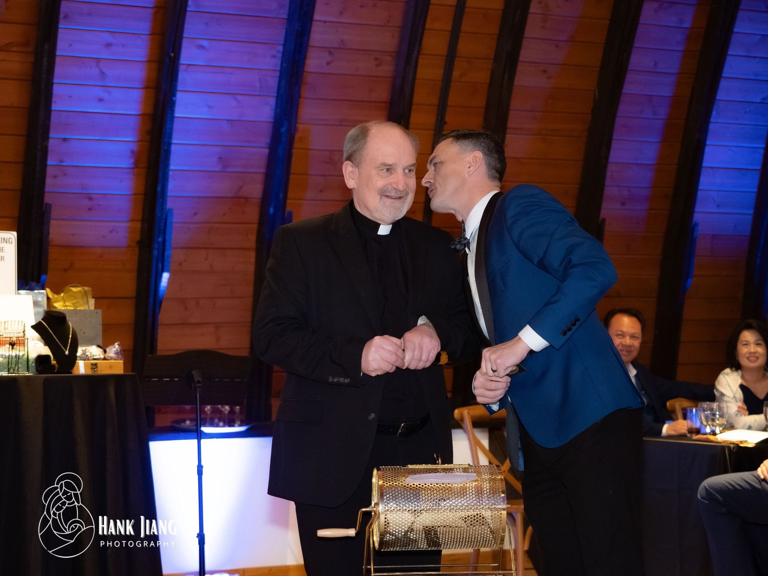A priest in black clergy attire shares a warm moment with a man in a suit at an indoor event.