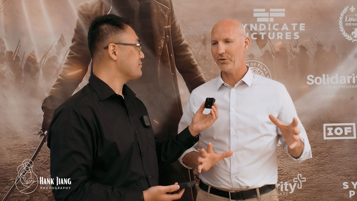 Two men in conversation at a Syndicate Pictures event, one in black shirt, other in white, discussing film industry topics.