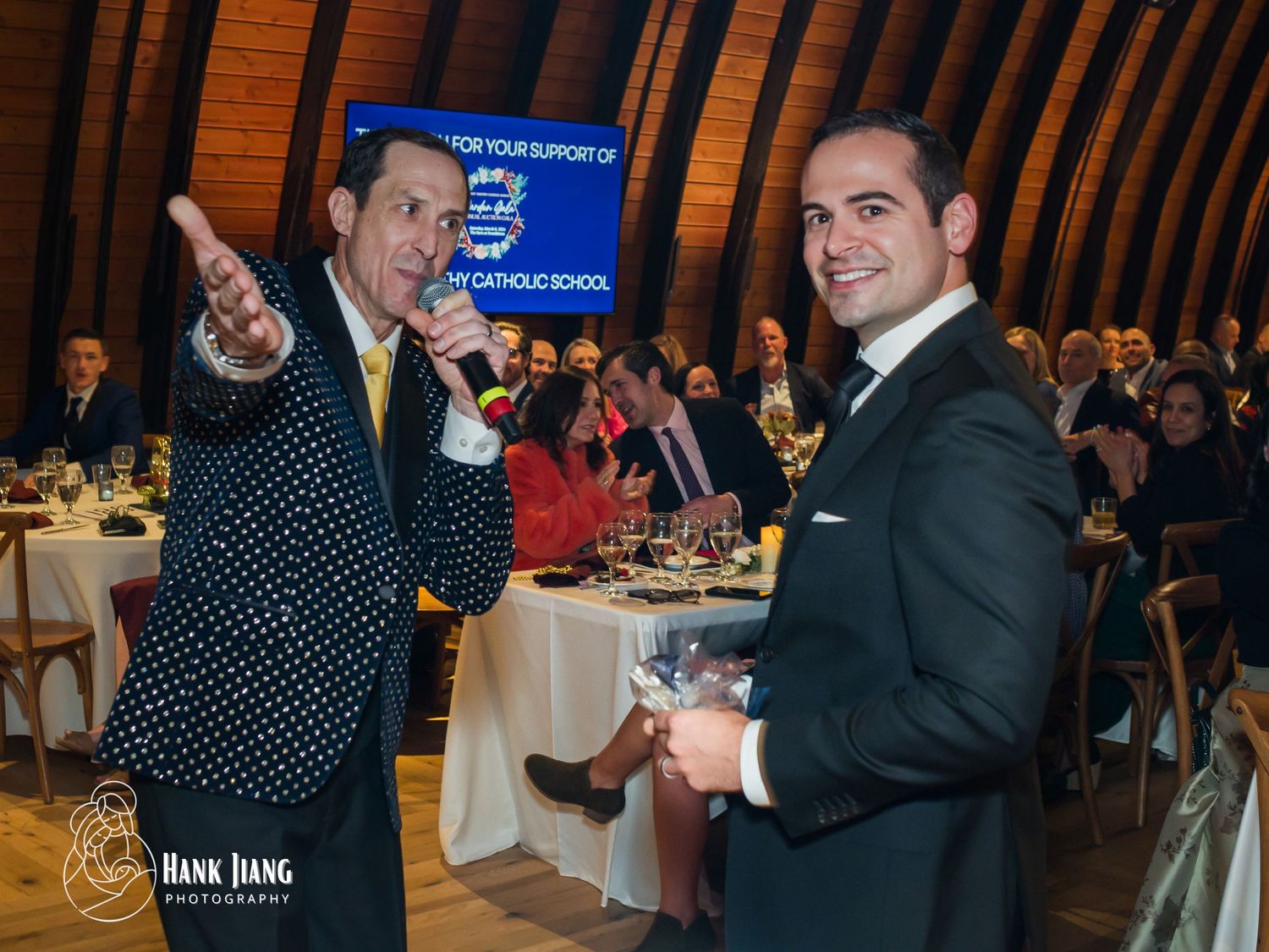 Two men in formal attire smile at a Catholic school fundraiser gala dinner event indoors.