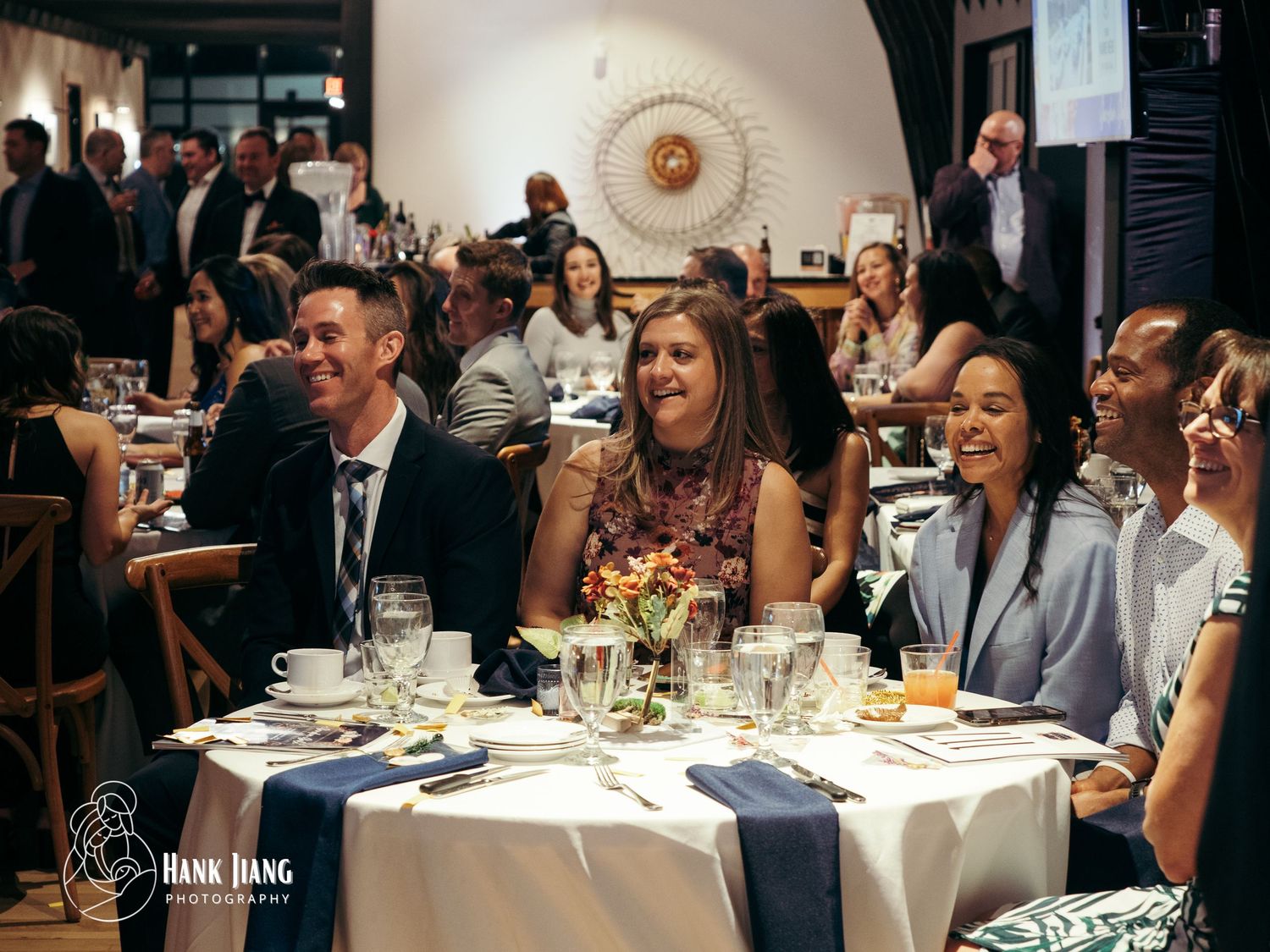 Guests laughing and smiling at a formal dinner event, seated at elegantly set tables in a stylish restaurant venue.