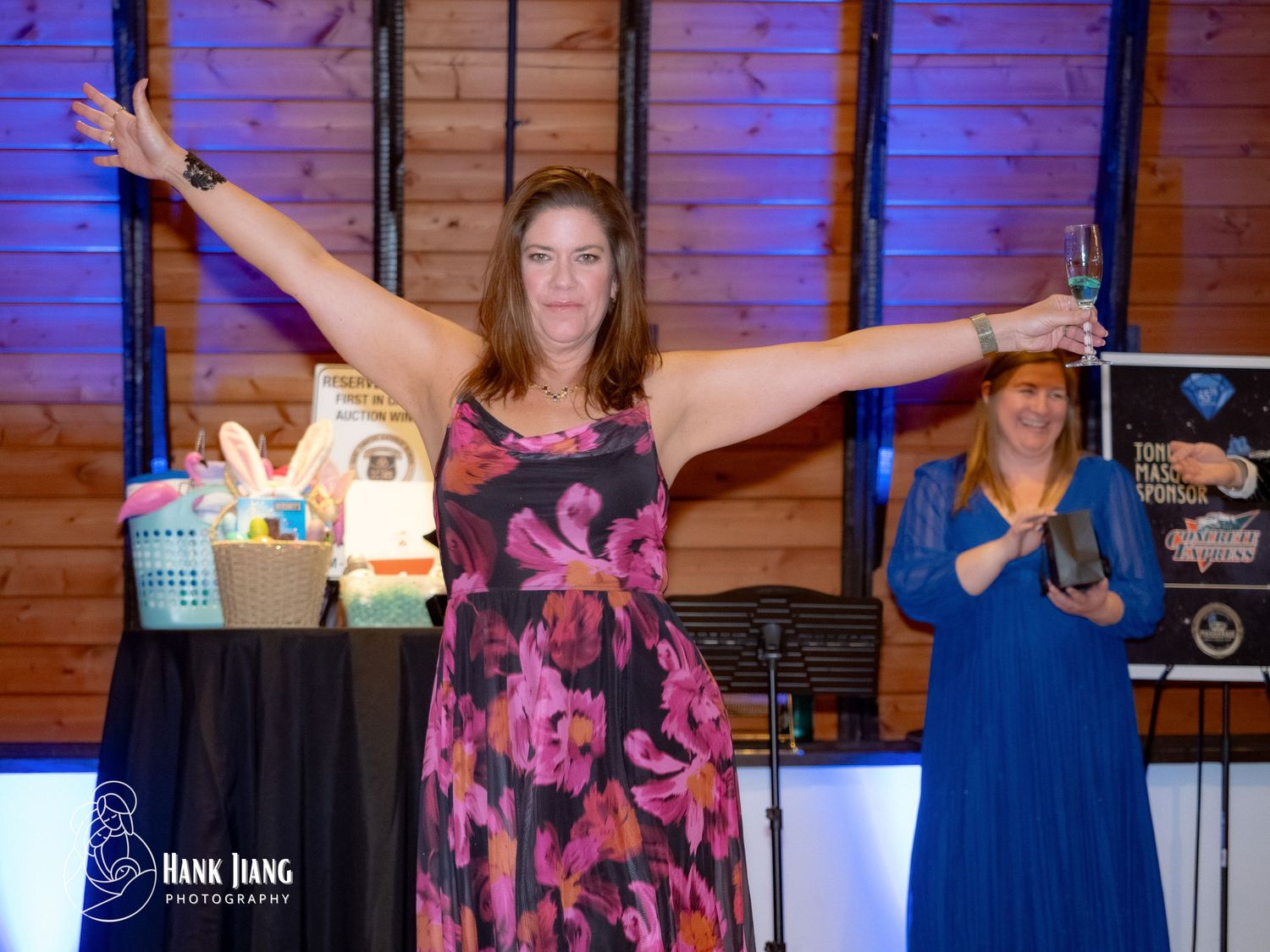 Woman in floral dress celebrates with arms outstretched at an indoor gala event with prizes on display.