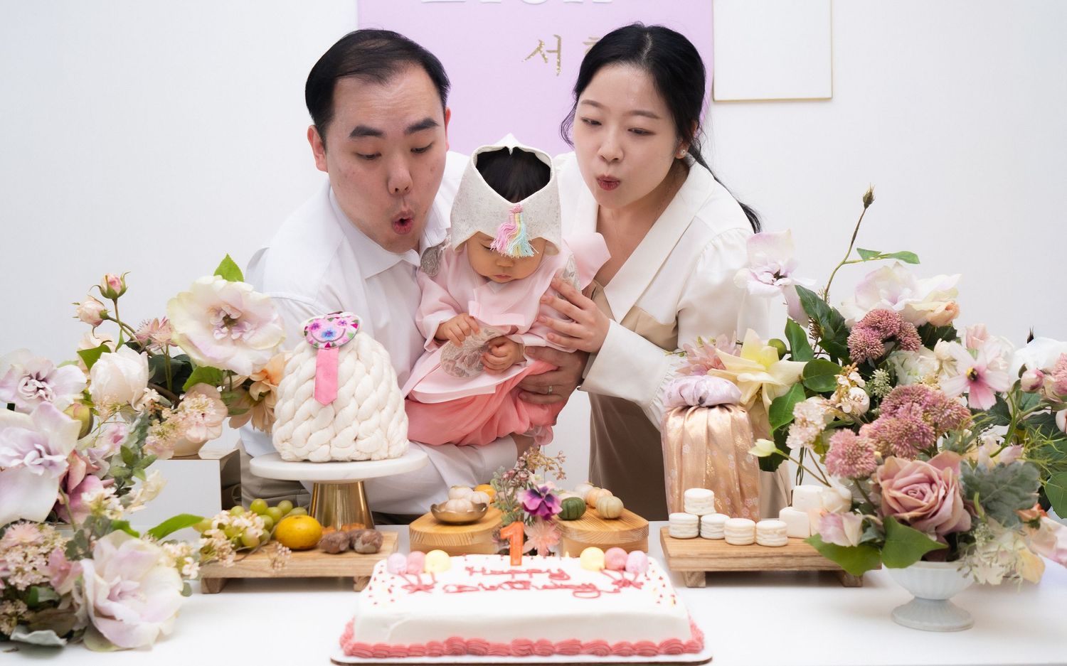 Korean family celebrates baby first birthday doljabi with parents blowing out candles surrounded by flowers and traditional foods.