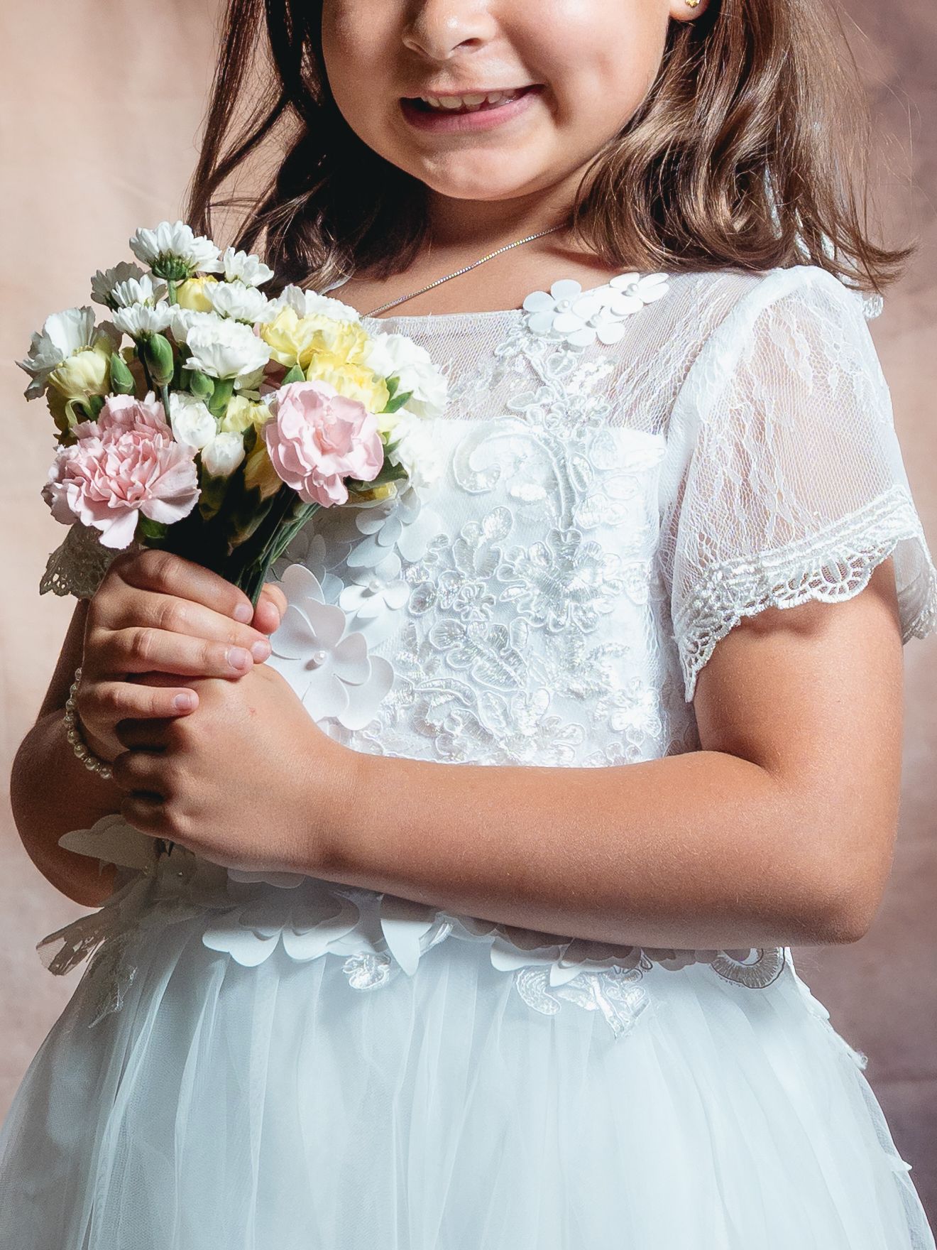 Young flower girl in white lace dress holding a bouquet of pink and yellow flowers, smiling at a wedding.