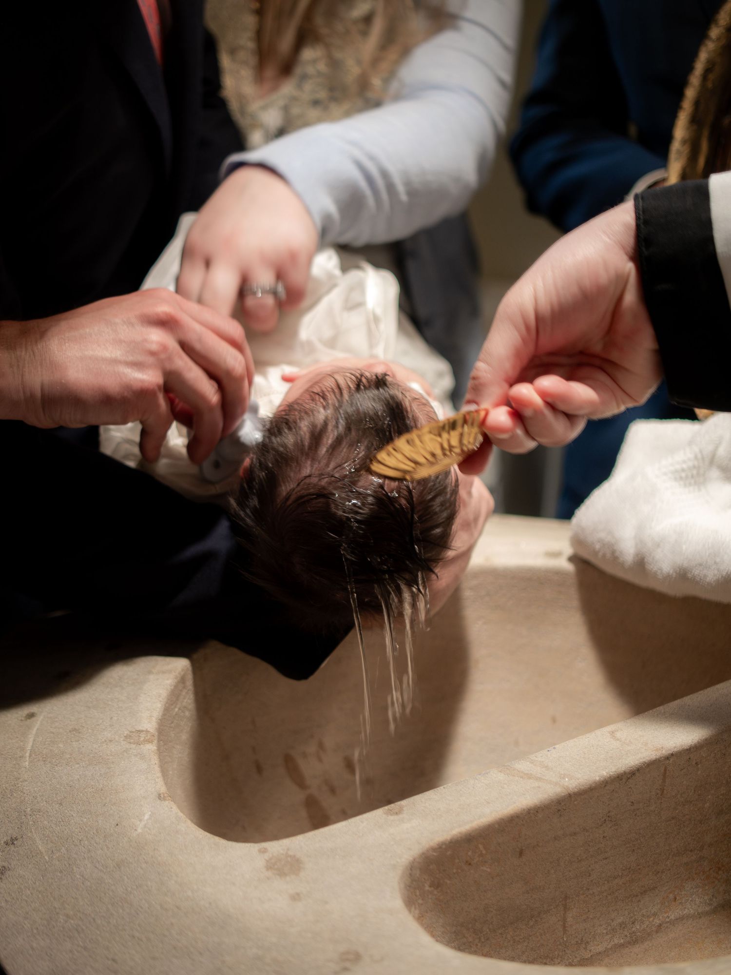 Baby being baptized with water poured over head at a stone baptismal font during a Catholic ceremony.
