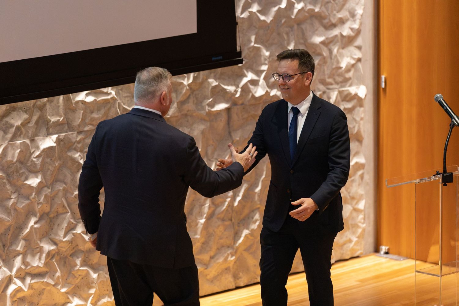 Two men in suits shaking hands on stage at a formal award ceremony in a modern venue.