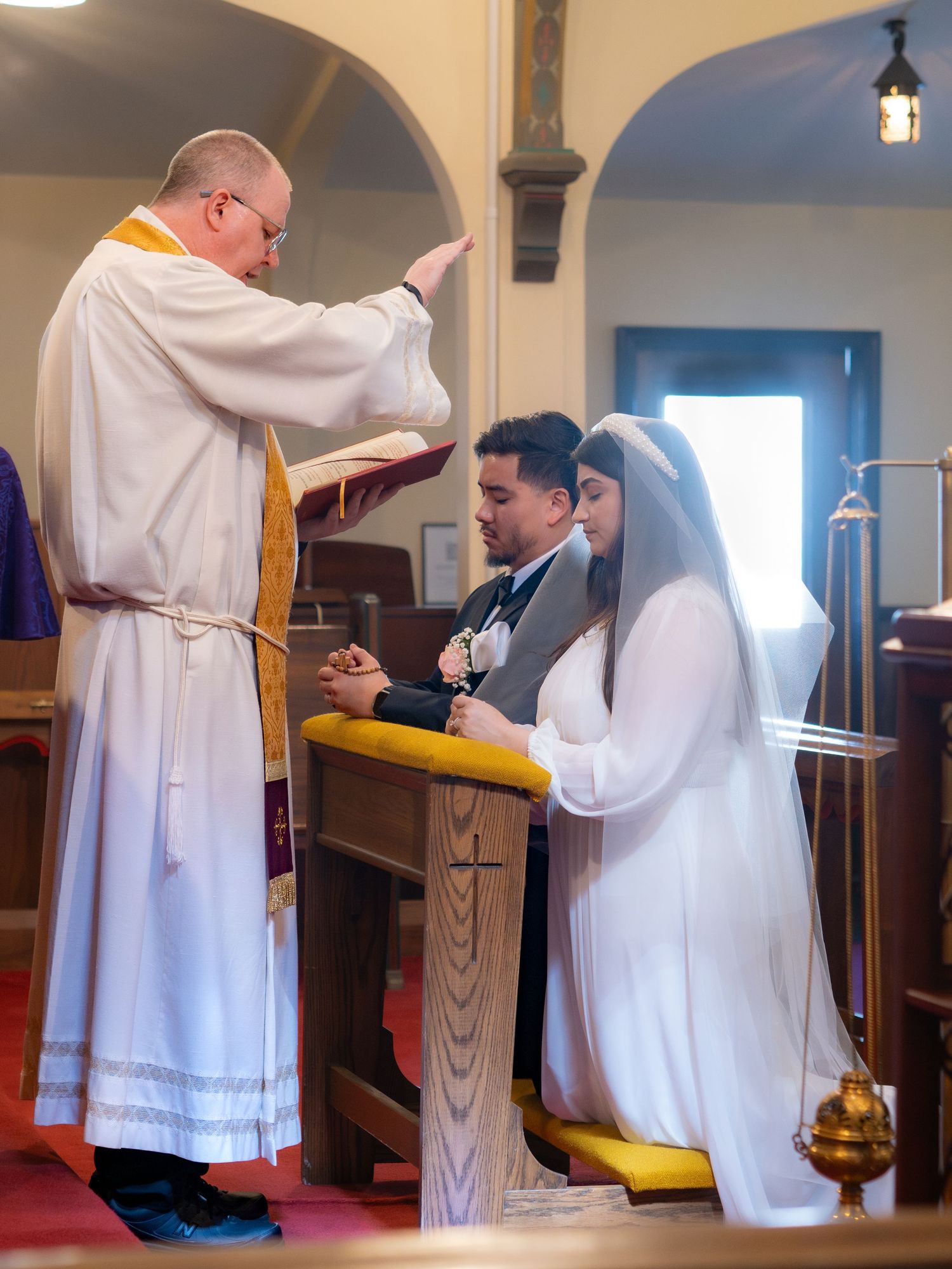 A priest blesses a kneeling bride and groom at a wooden altar during a church wedding ceremony.