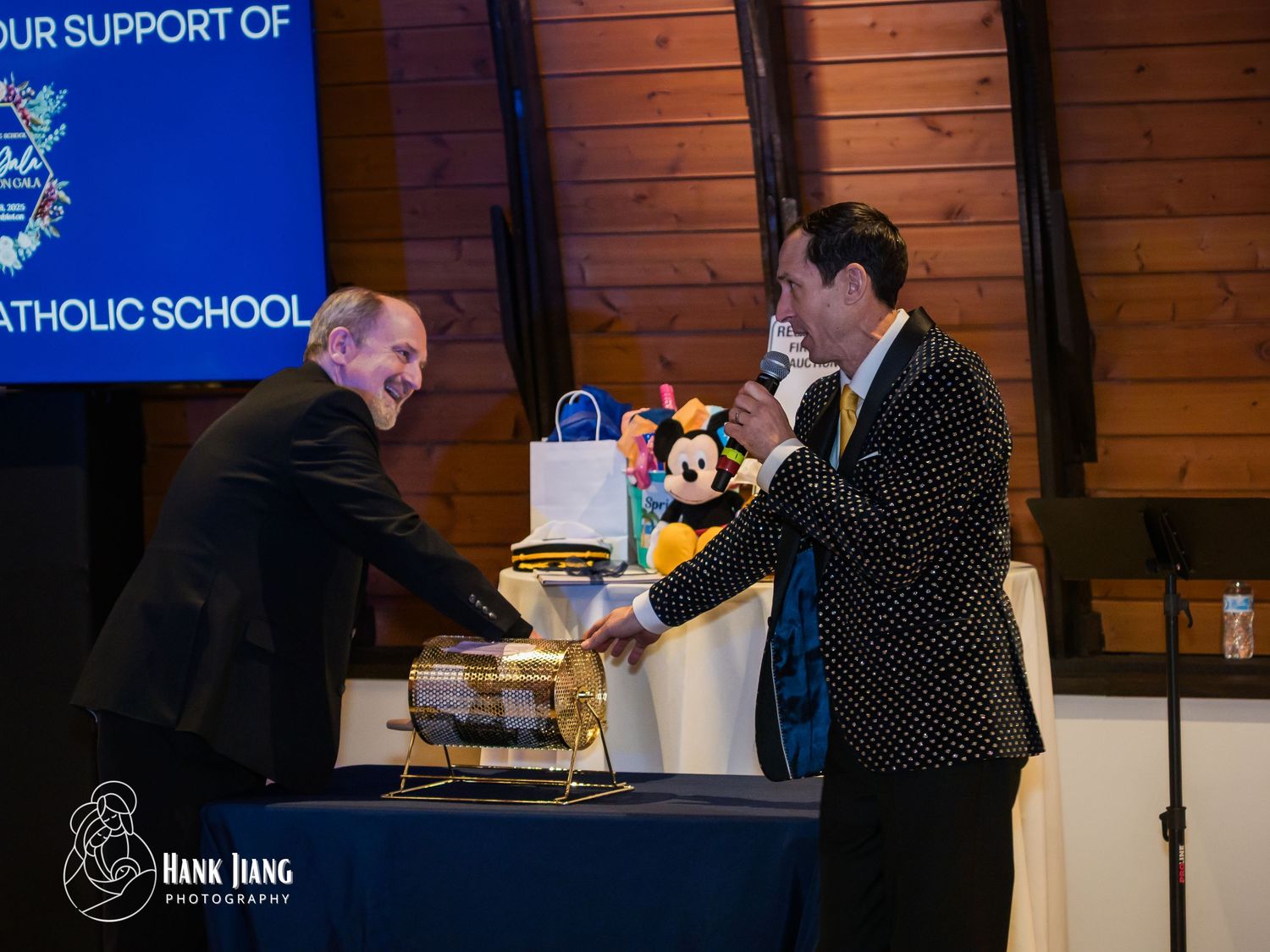 Two men shake hands at a Catholic school fundraiser event with Mickey Mouse plush and gifts on display.