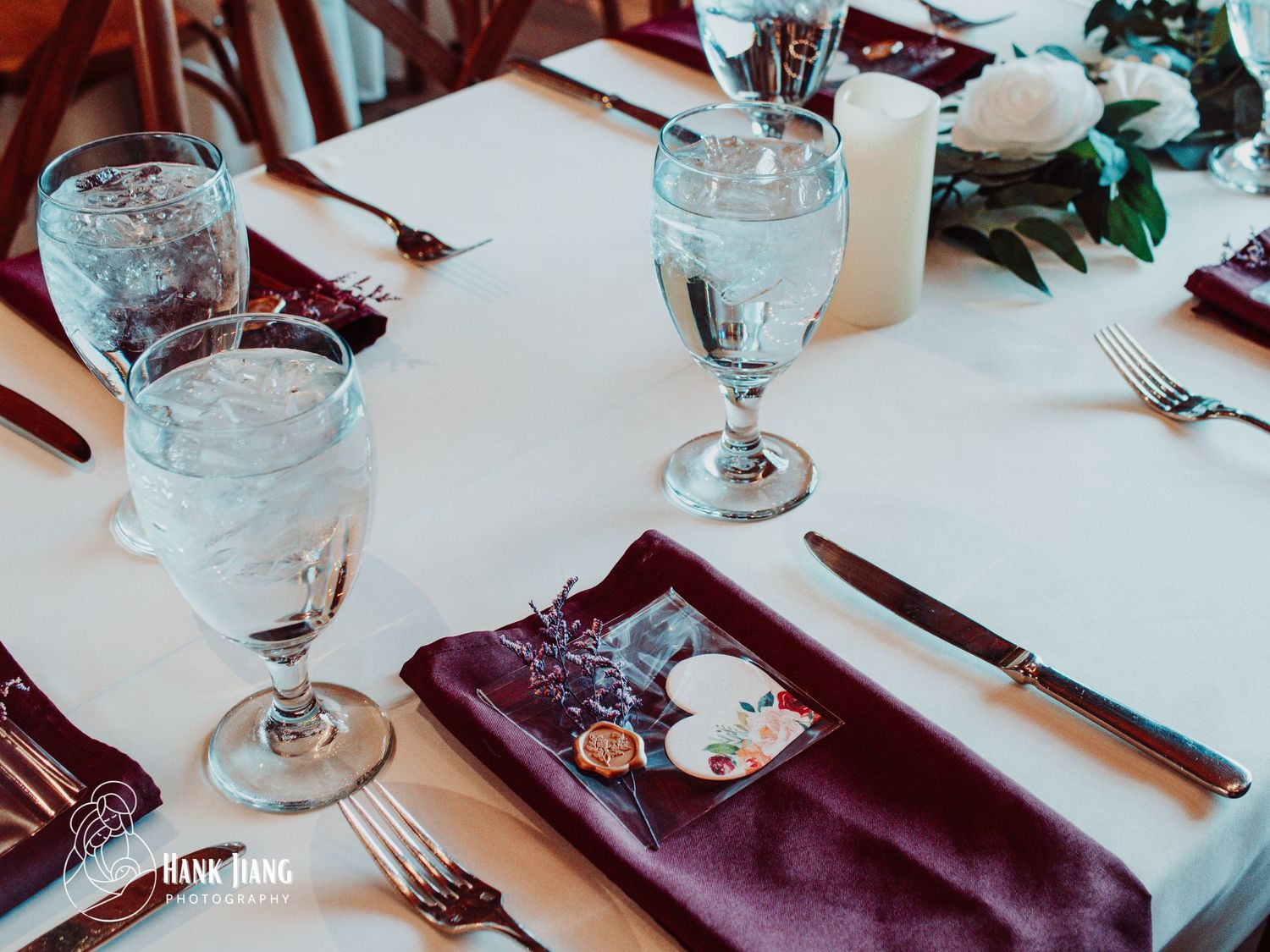 Elegant wedding reception table setting with burgundy napkins, crystal glasses filled with water, and floral centerpiece.