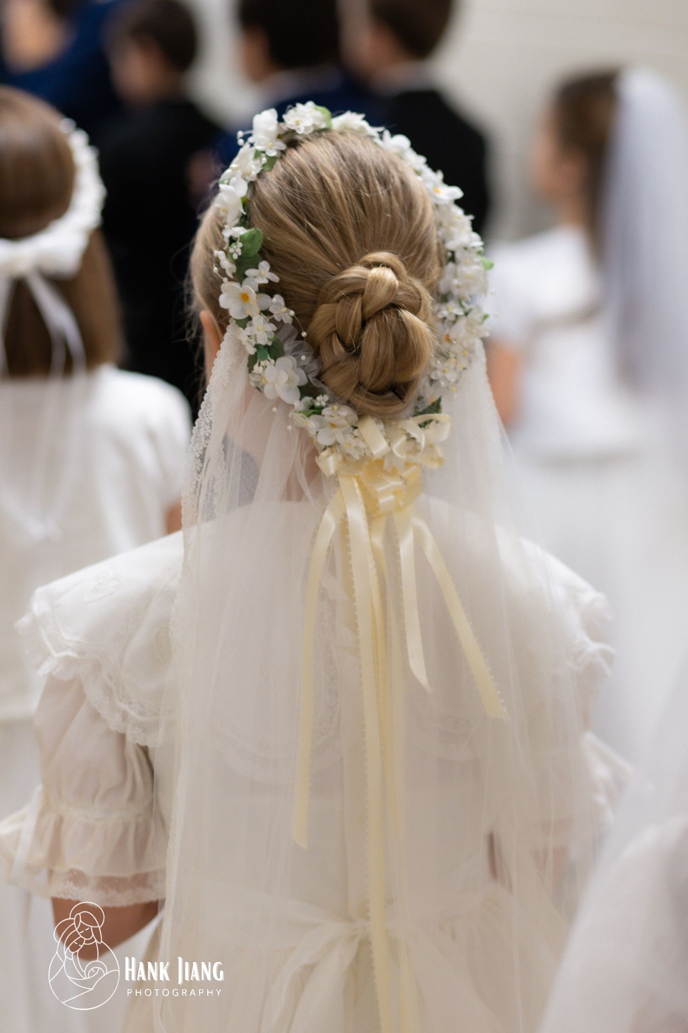 Girl with braided updo adorned with white flower crown and veil with yellow ribbon at First Communion ceremony.