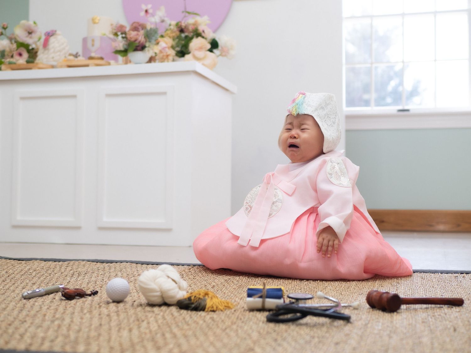 Baby in pink Korean hanbok crying during Dol first birthday celebration with traditional items on floor.
