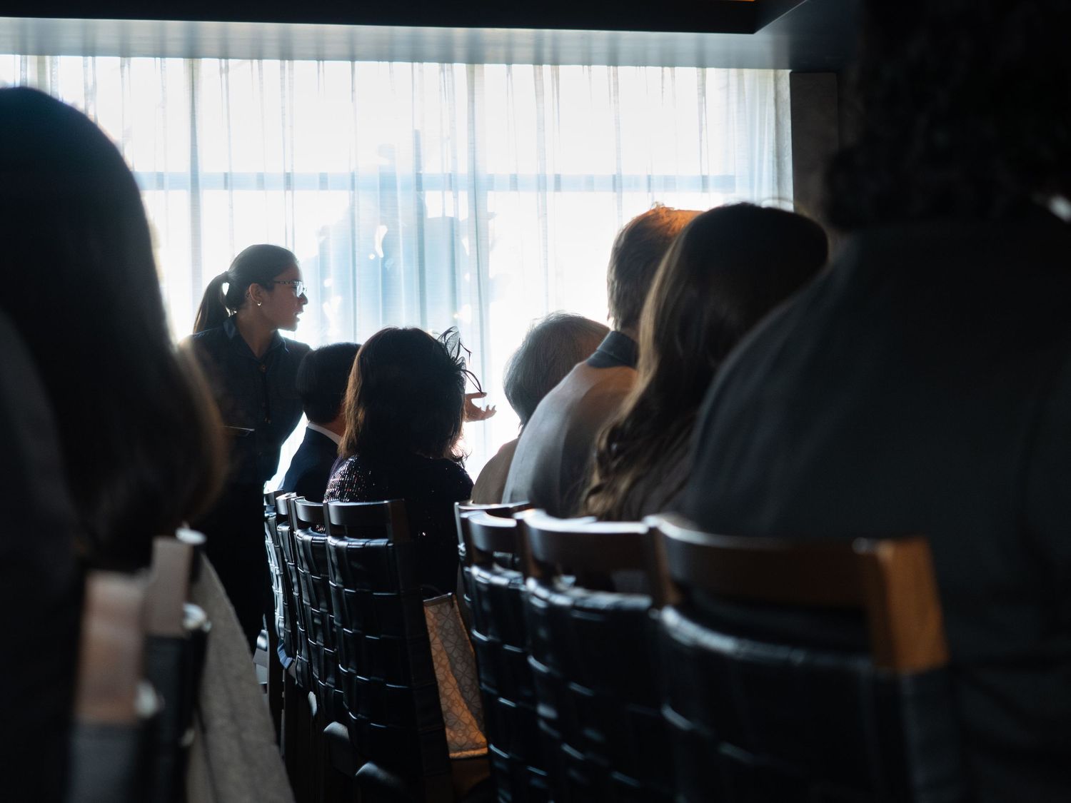 People seated in rows of chairs at an indoor event, with soft backlit window light creating a moody atmosphere.