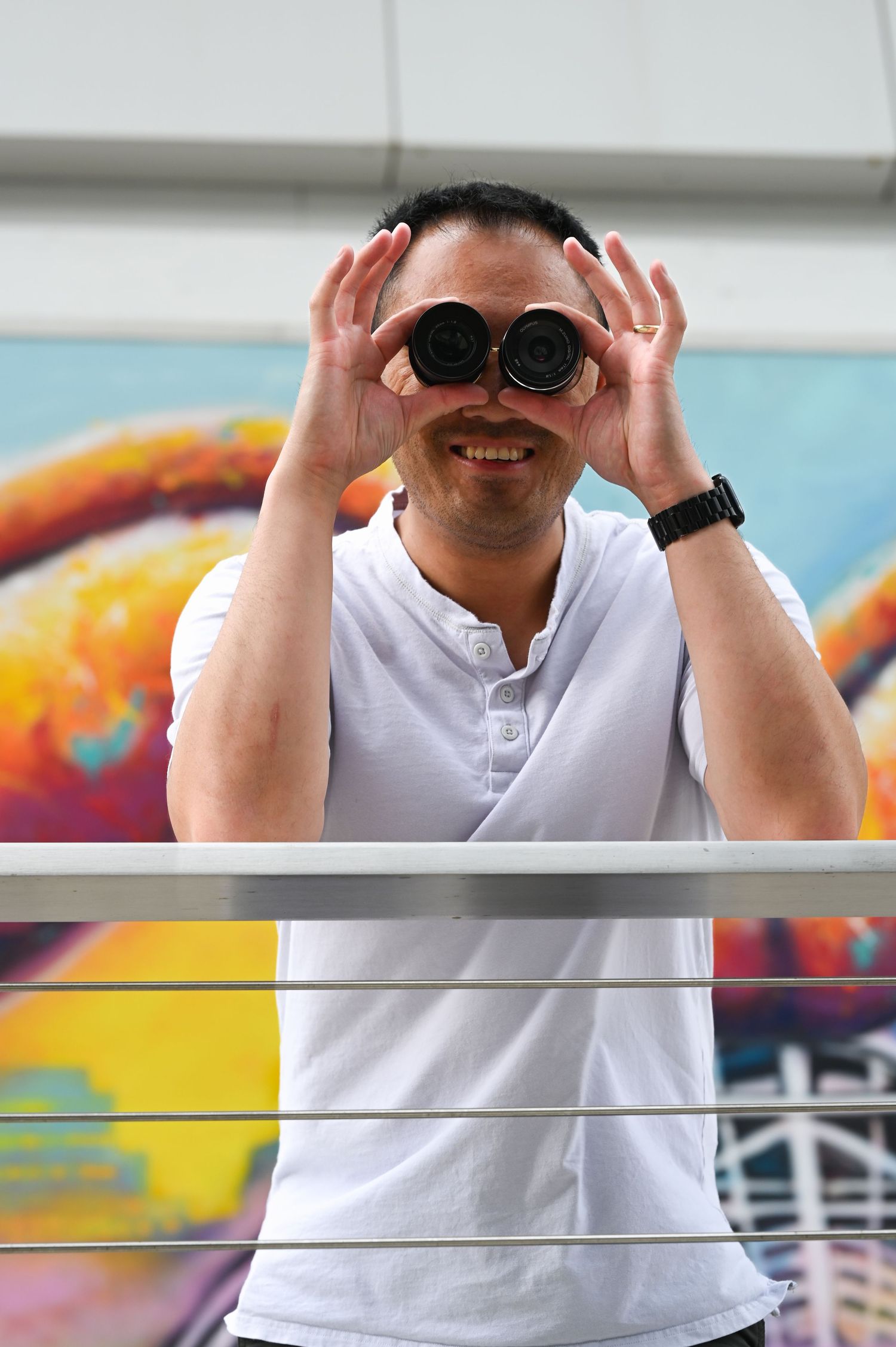 Hank Jiang, a male photographer, is humorously holding two camera lenses over his eyes like binoculars outdoors.