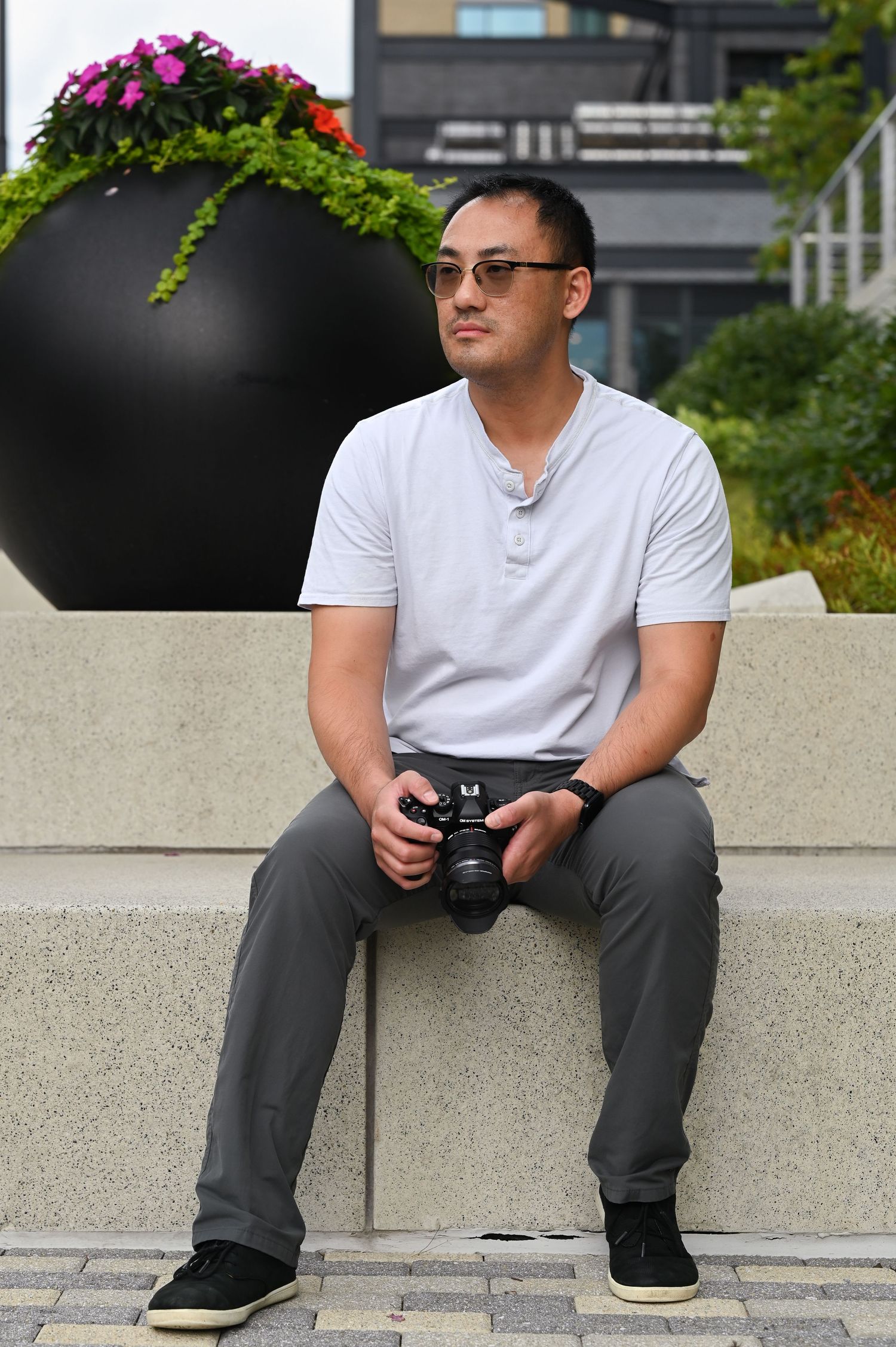 Hank Jiang, a male photographer, is sitting on a stone bench holding mirrorless camera in urban outdoor setting.