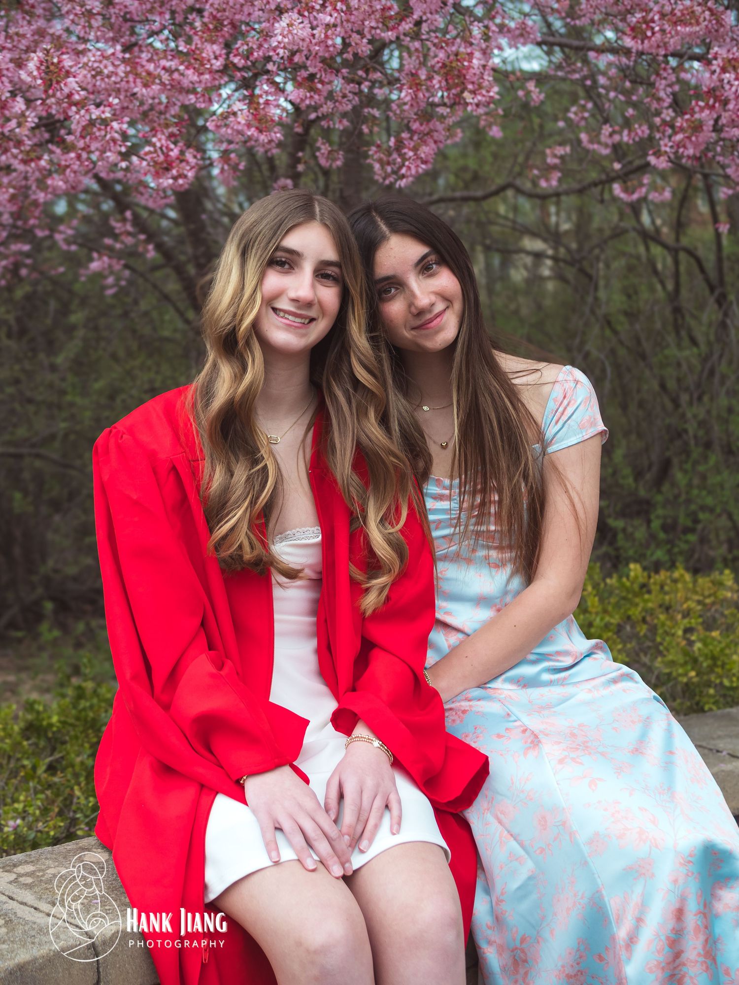 Two young women in red and blue dresses pose together under blooming pink cherry blossom trees in spring.