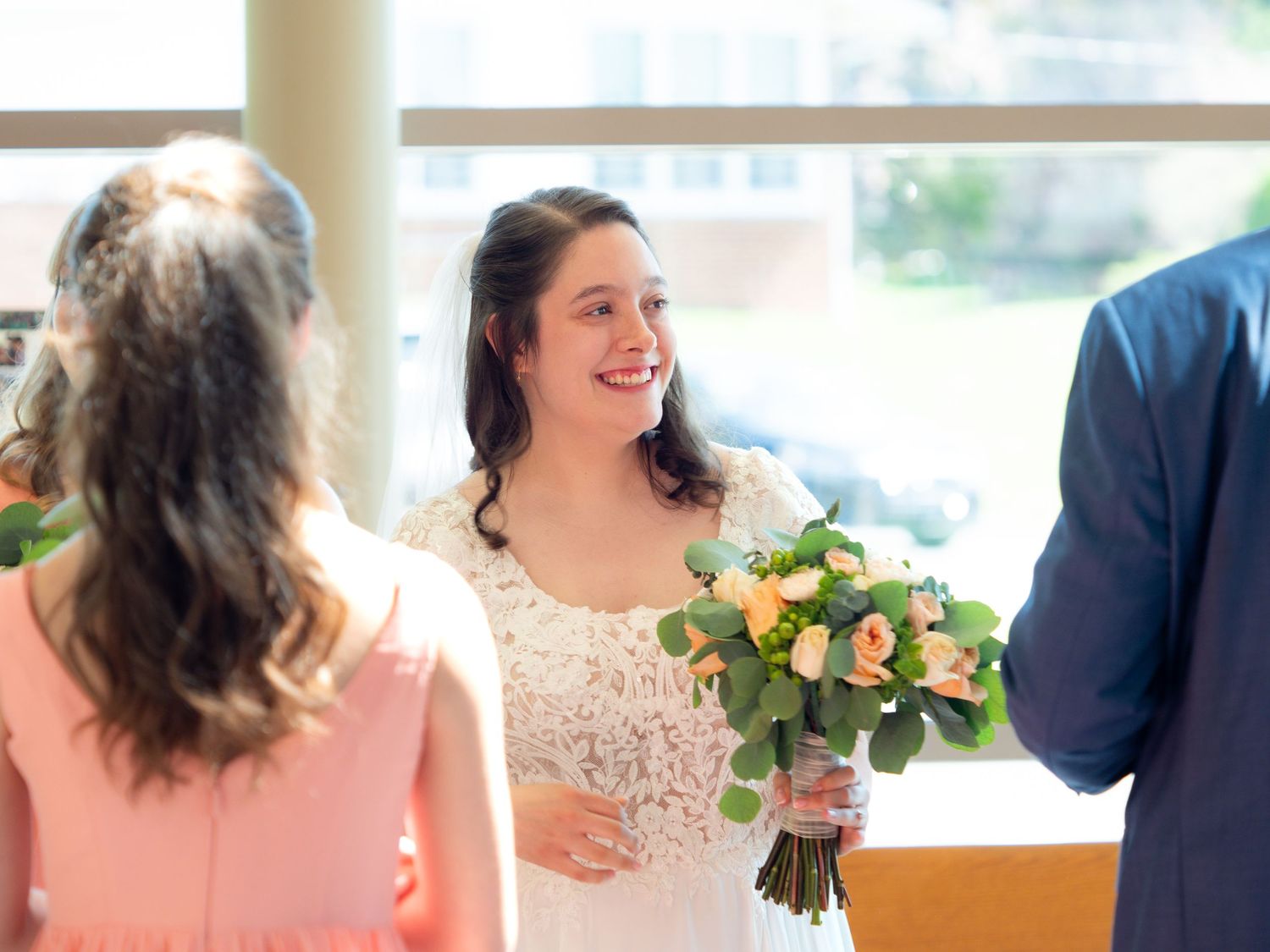 A smiling bride in a lace gown holds a peach and green bouquet while greeting guests at her wedding.