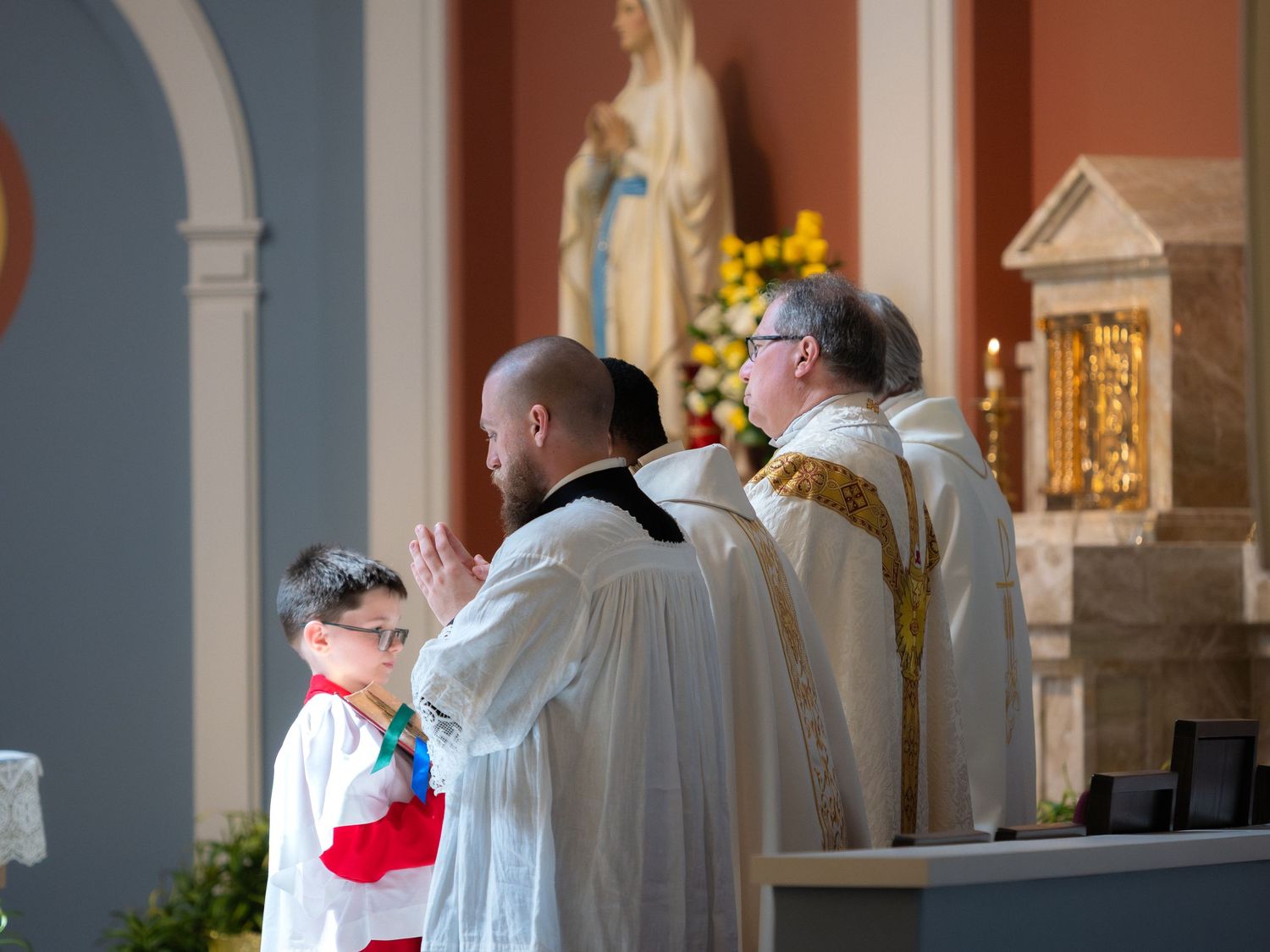 Catholic Mass ceremony with altar servers and priests praying before a statue of Mary in a church.