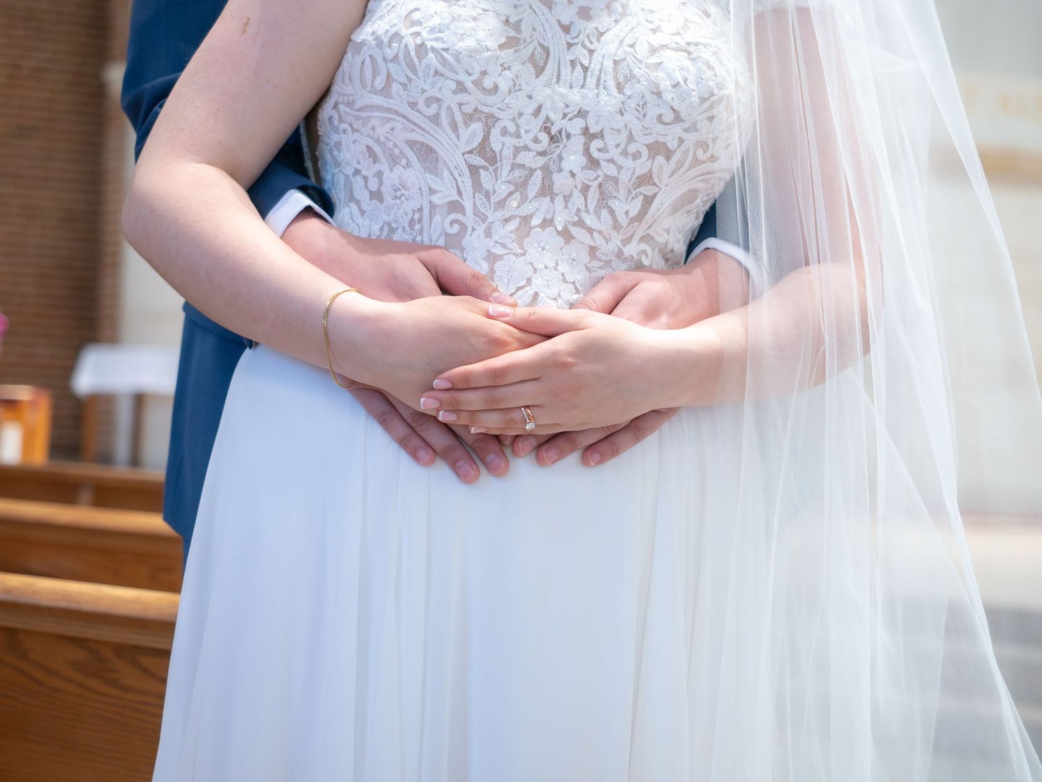 Groom embracing bride from behind, both with hands clasped at her waist in church.