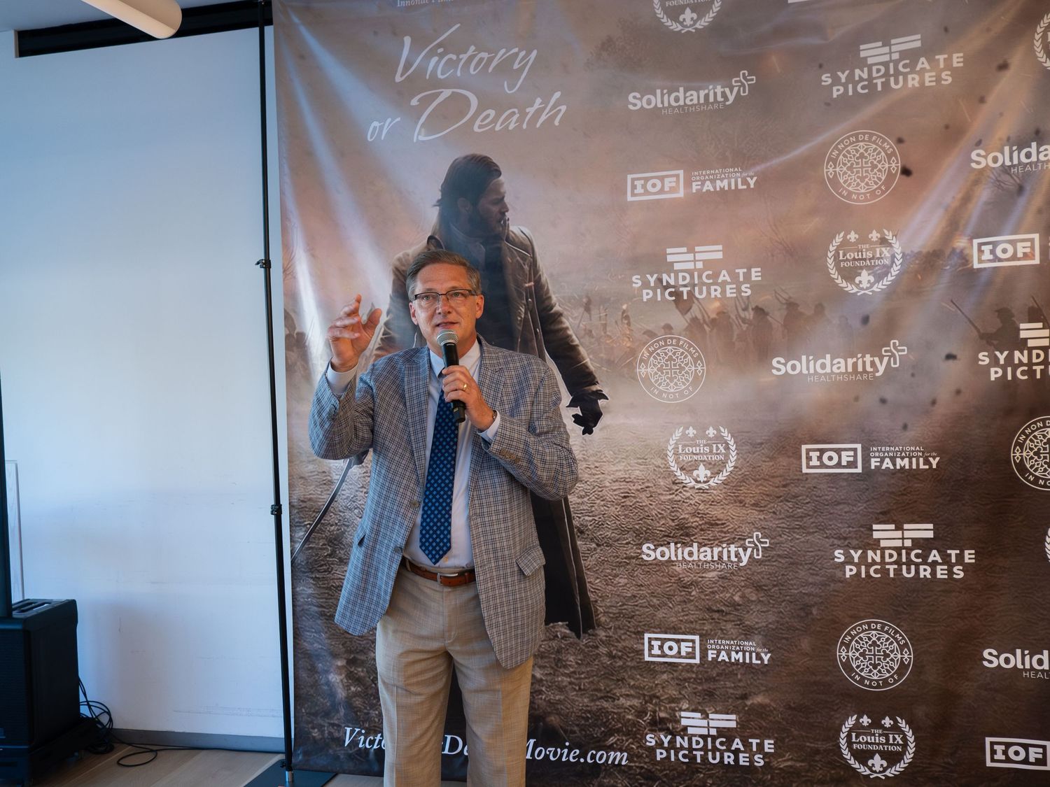 Man speaking at Victory or Death film premiere event, gesturing confidently before a branded step-and-repeat backdrop.