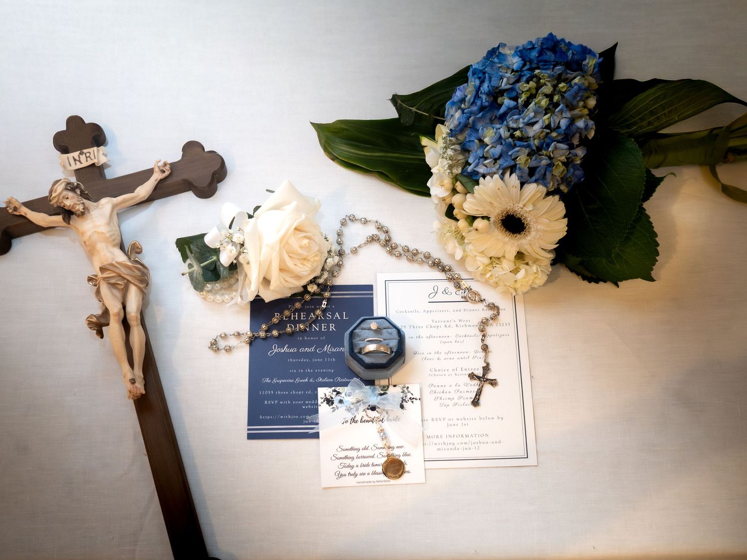 Catholic wedding flat lay with rosary, rings, cross, blue hydrangeas, and white roses on a white surface.