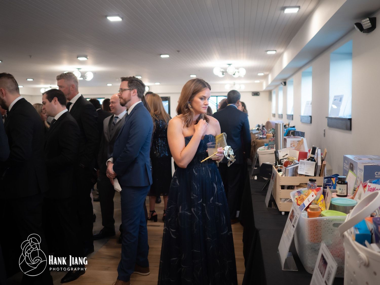 Elegantly dressed guests mingle at a formal gala event, with a woman in a navy gown browsing a display table.