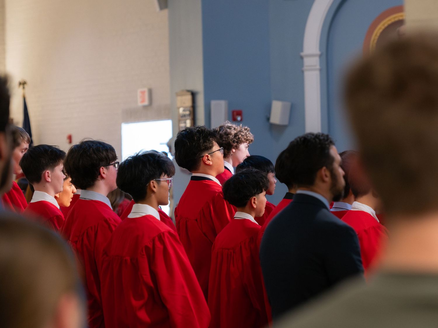 Students in red graduation gowns stand in rows during a formal ceremony inside a blue-walled hall.